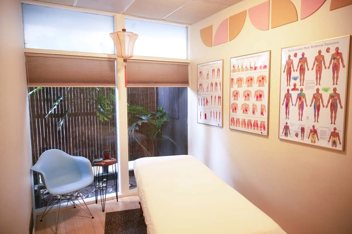 A small room with a massage table, a white chair, and a small side table. There are four anatomical acupuncture posters on the wall and a window with closed blinds, overlooking a garden.