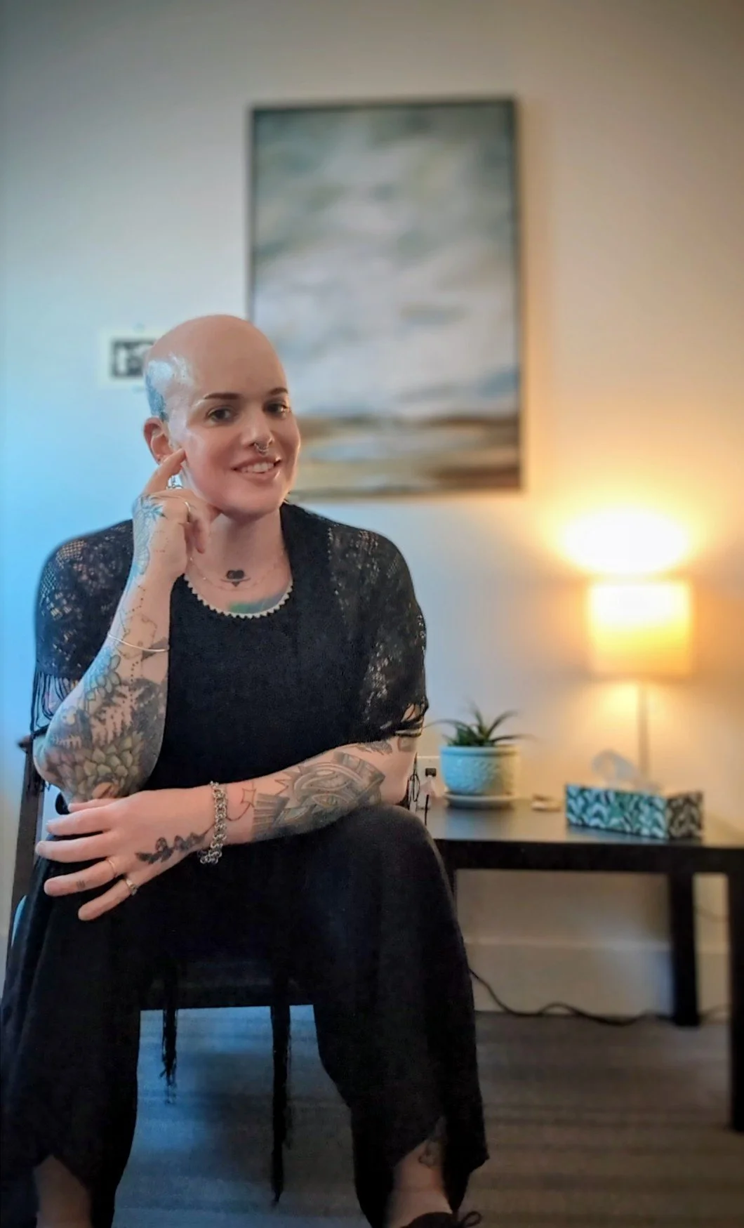 A woman with tattoos and a shaved head smiling and sitting on a chair in a living room with a painting, a lamp, and a potted plant in the background.