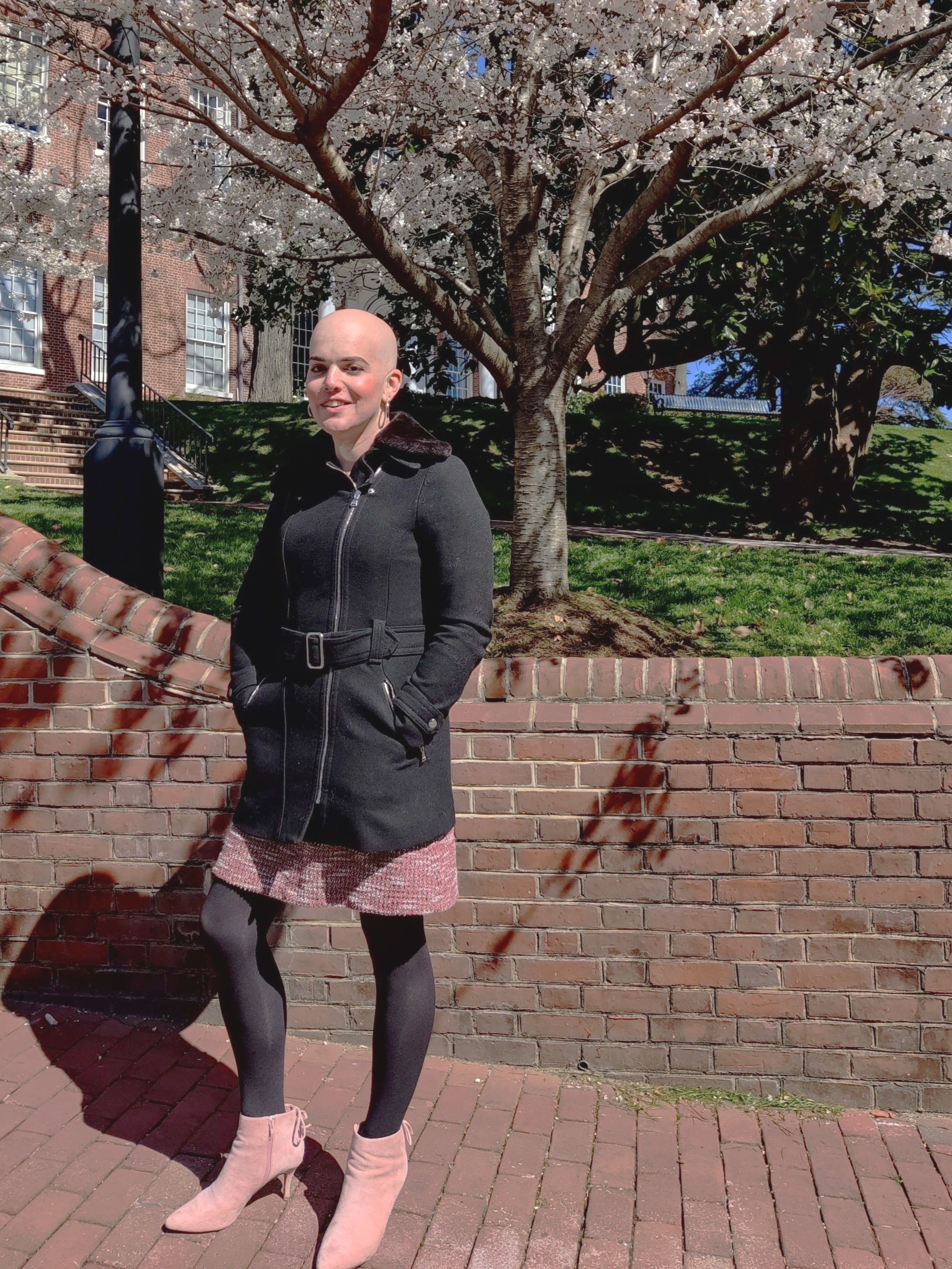 A woman stands outdoors on a brick sidewalk near a brick wall, with a blooming tree and grassy area in the background. She is wearing a black coat, black tights, beige heeled boots, and has a bald head. She is smiling and has one hand in her coat pocket.