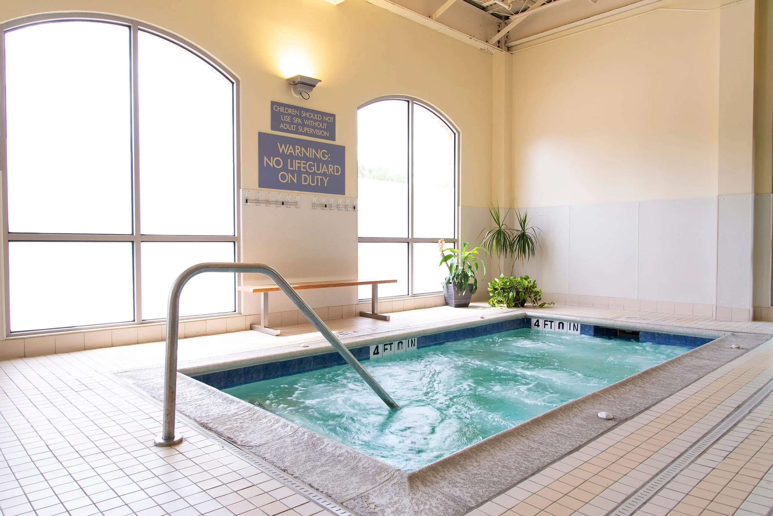 Indoor hot tub with metal handrail, large windows, potted plants, blue and white signs with safety instructions, and tiled floor.