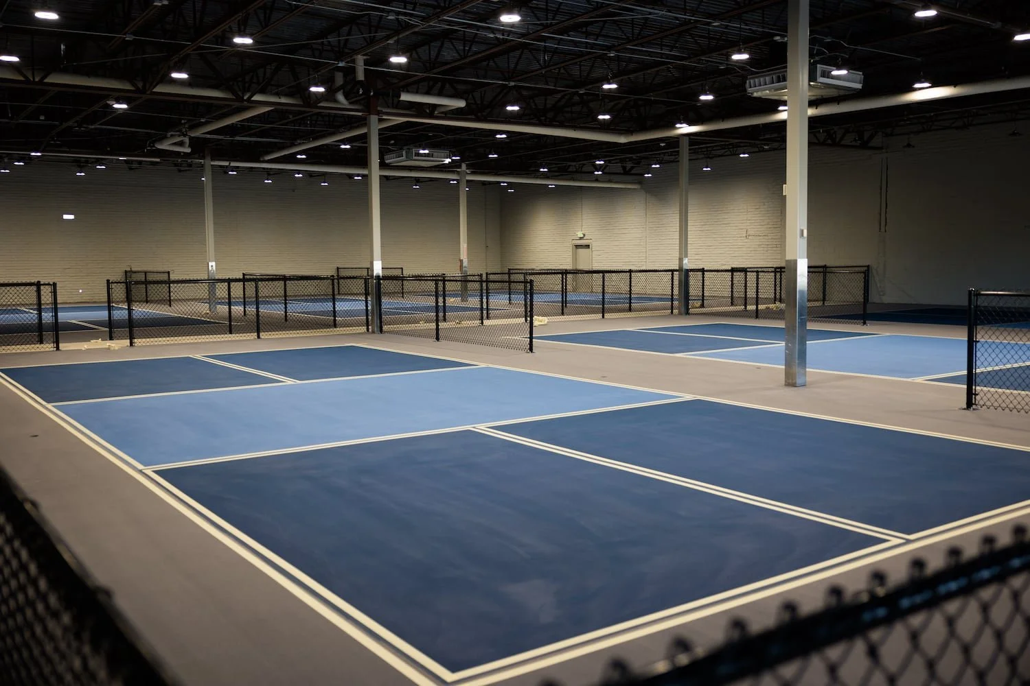 Indoor tennis courts with blue playing surfaces, surrounded by black chain-link fences, under a high ceiling with lights.