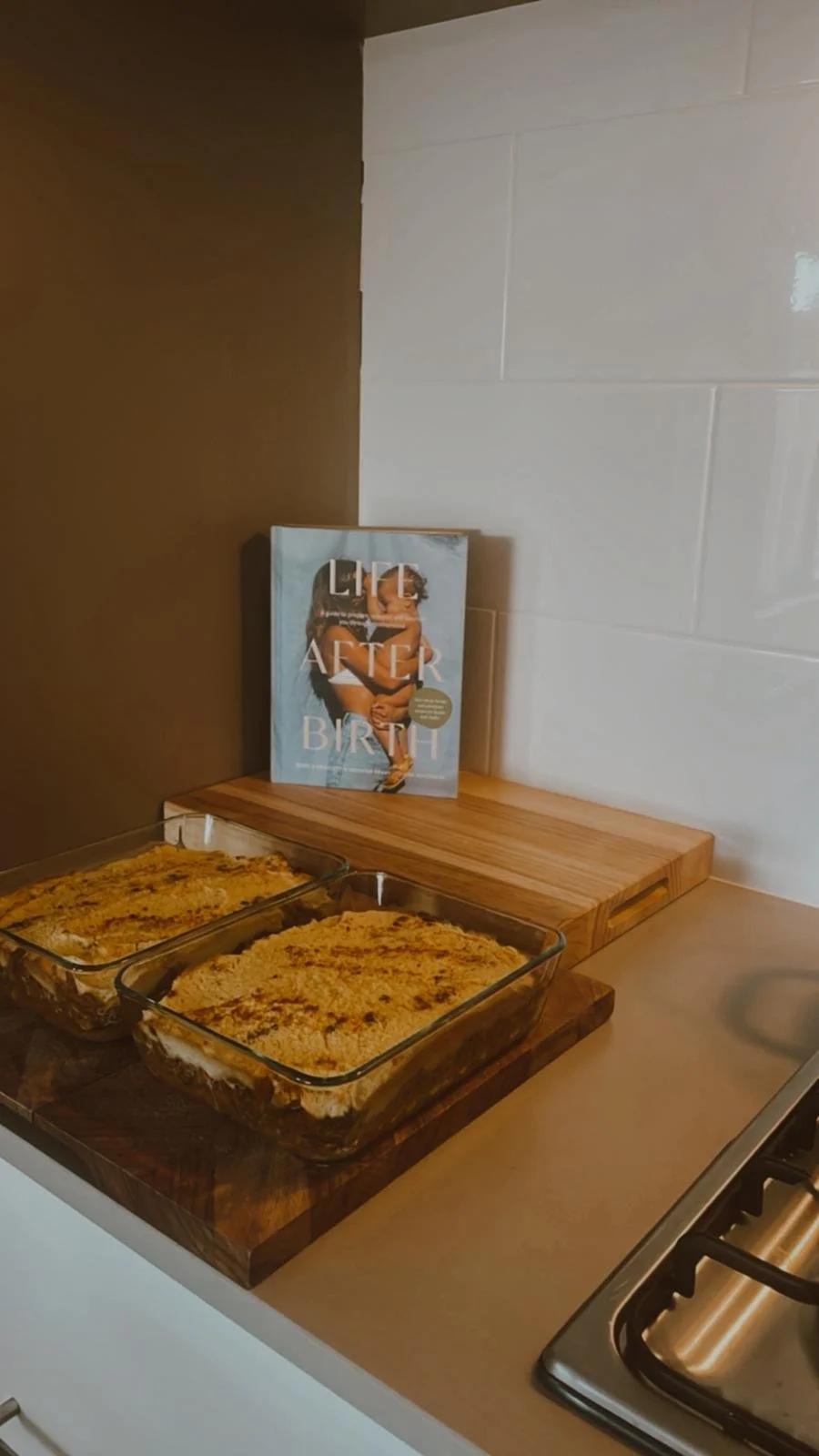 Two baking dishes with lasagna on a kitchen counter, a cookbook titled 'Life After Birth' standing behind them, and a wooden cutting board beside the cookbook.