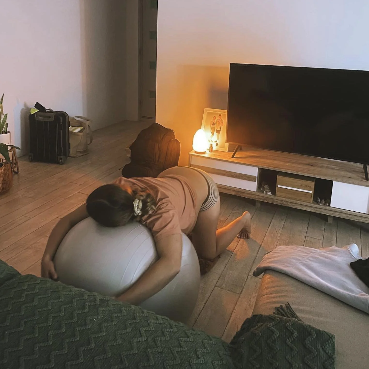 A woman is kneeling on a living room floor, leaning over a large exercise ball, with her head and arms resting on it. The room has a television, a white and wood TV stand, a salt lamp, framed photo, and other decorative items. The lighting is warm, creating a cozy atmosphere.