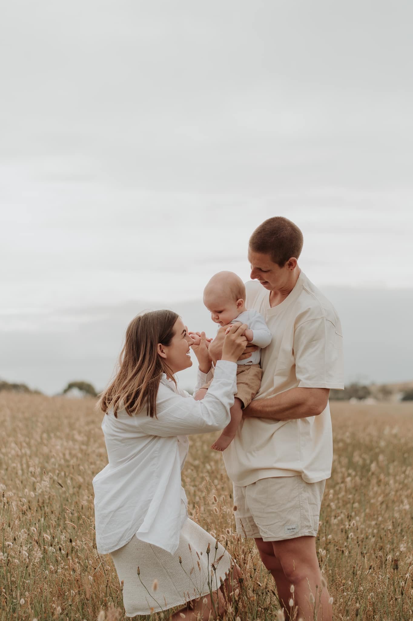 A family of three outdoors in a field of tall grass, with the mother kneeling and smiling at her baby, while the father holds the baby and looks at the mother, under a cloudy sky.