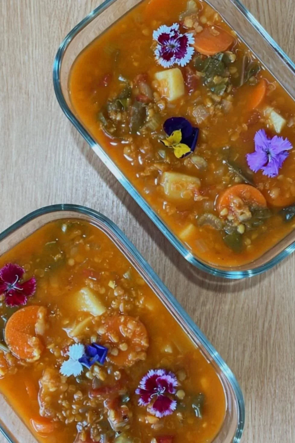 Two glass baking dishes filled with vegetable soup garnished with edible flowers on a wooden surface.