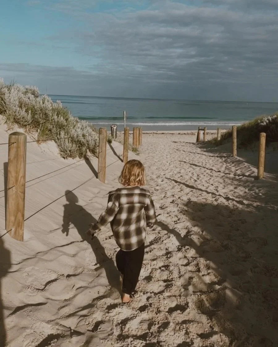 A person with curly blonde hair walking barefoot on a sandy beach towards the ocean, with a cloudy sky overhead and dunes on either side.