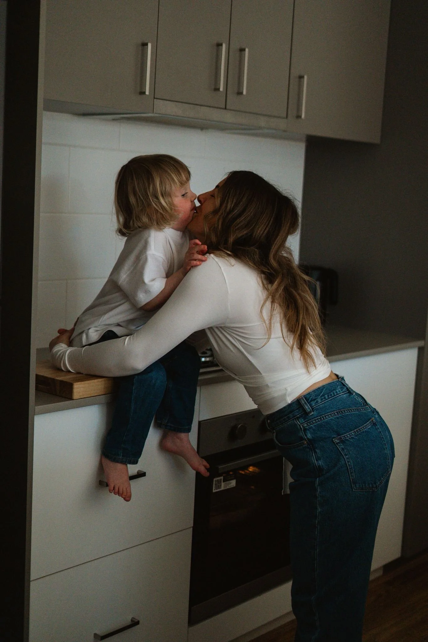 A woman and a young boy sharing a kiss in a kitchen, with the boy sitting on the counter and the woman leaning in to kiss him.