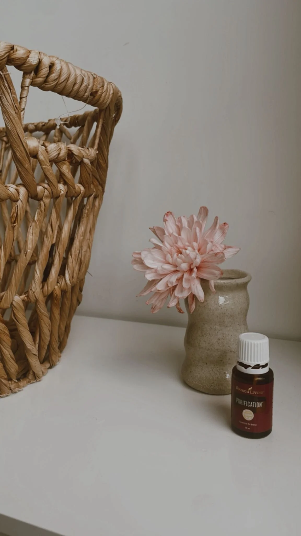 A wicker basket, a pink flower in a beige ceramic vase, and a small bottle of Young Living Purification essential oil on a white surface.