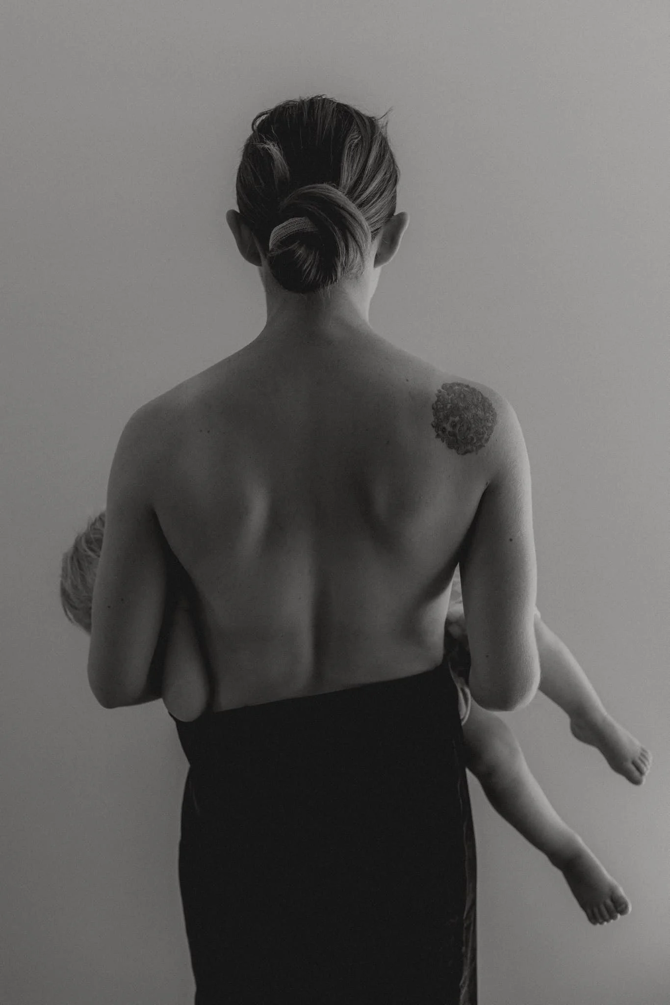 Black and white photo of a woman viewed from the back, holding a child in her arms. She has a tattoo on her right shoulder.