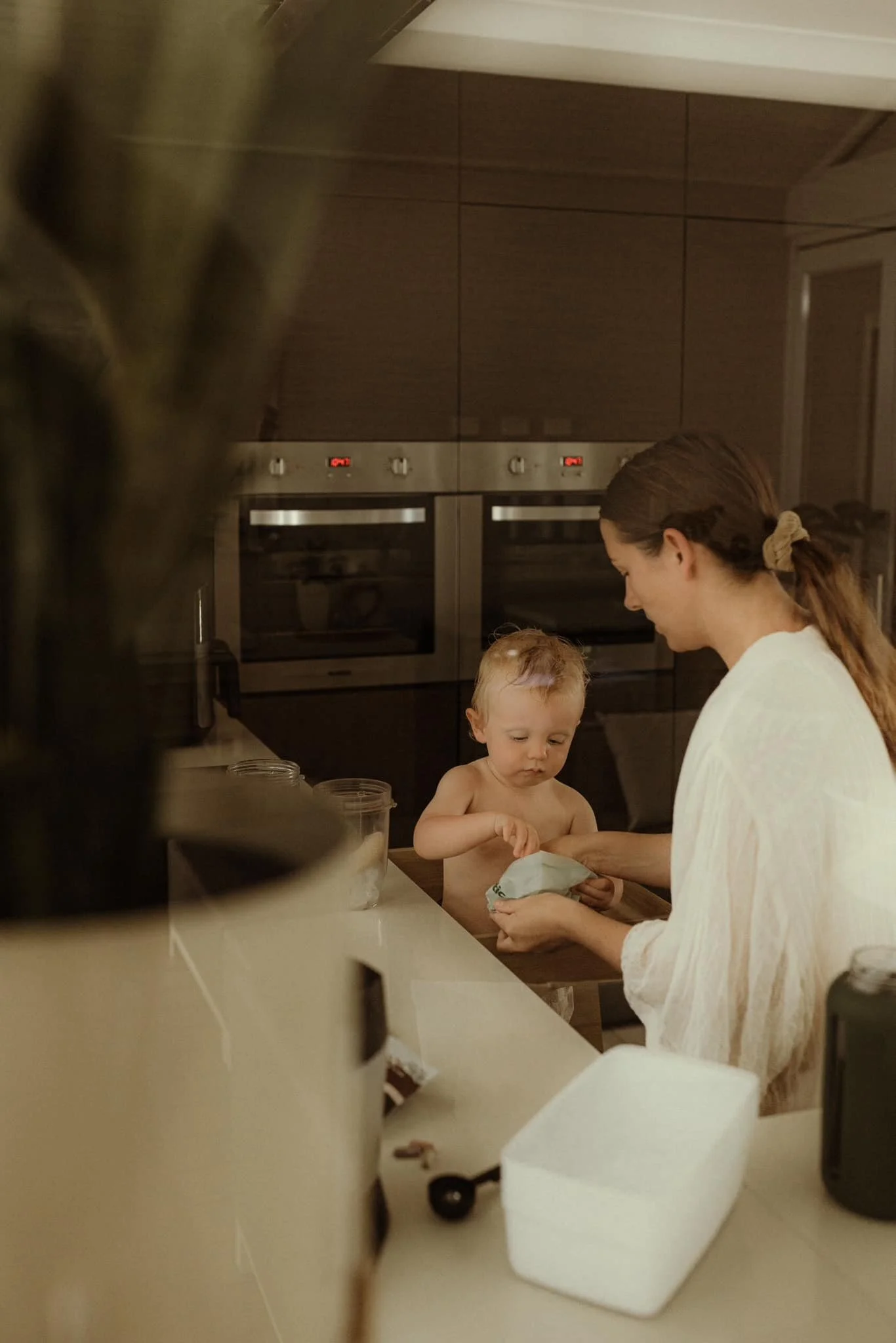 A woman and a young child with light skin and blonde hair are in a modern kitchen, with the woman handing something to the child. The child is shirtless, and they are near a kitchen counter with jars and a white container. The background features built-in ovens and dark wood cabinetry.