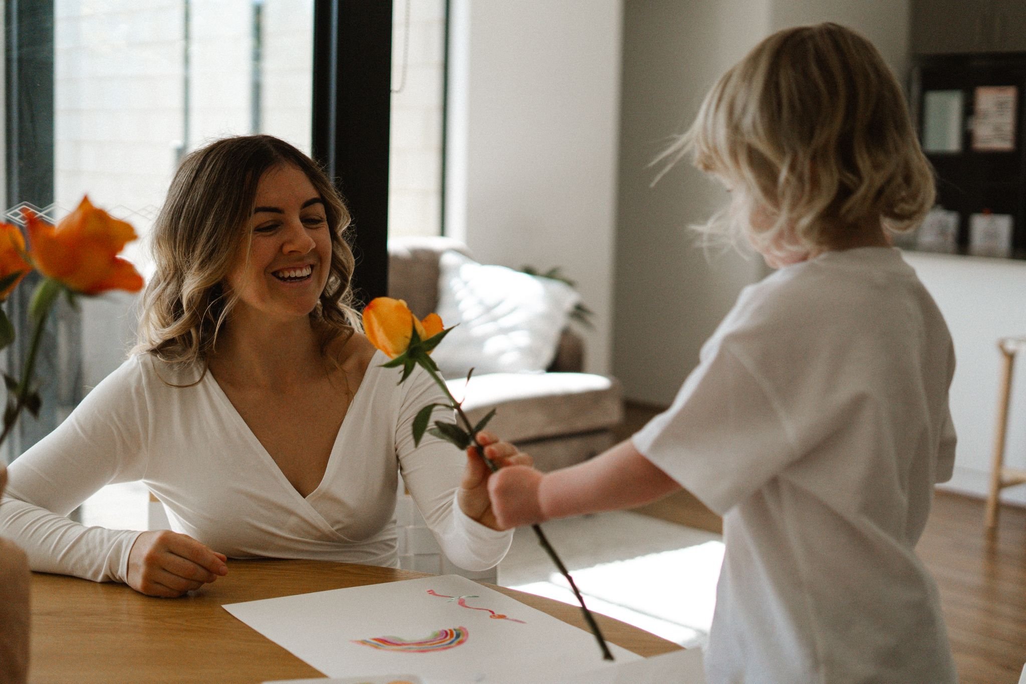 A woman is smiling and sitting at a table while a child gives her a yellow and orange flower.