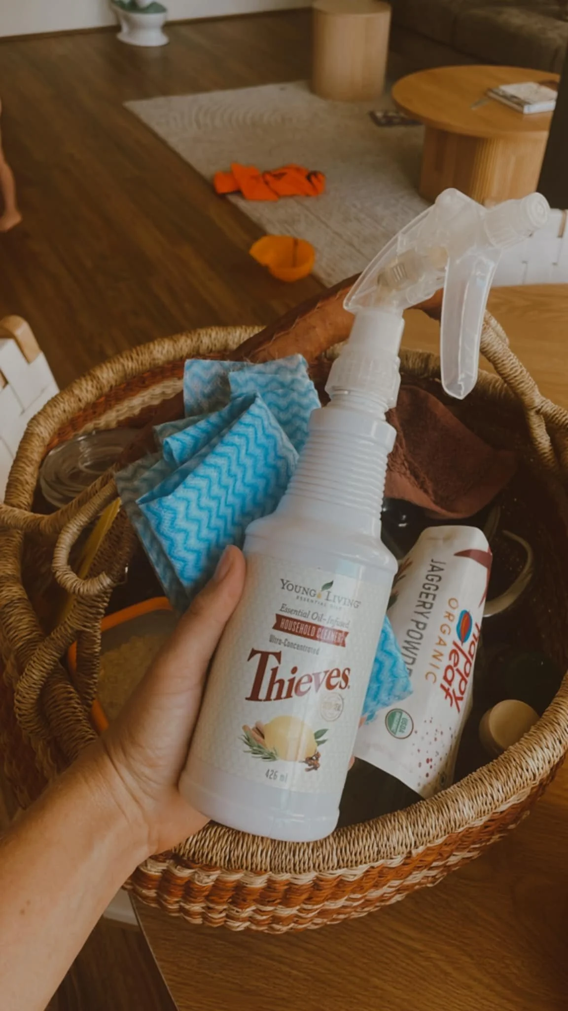 A hand holding a spray bottle labeled 'Thieves' with cleaning supplies in a wicker basket on a wooden table. In the background, there's a living room area with a rug, a small orange bowl, an orange cloth, and a wooden coffee table with books.