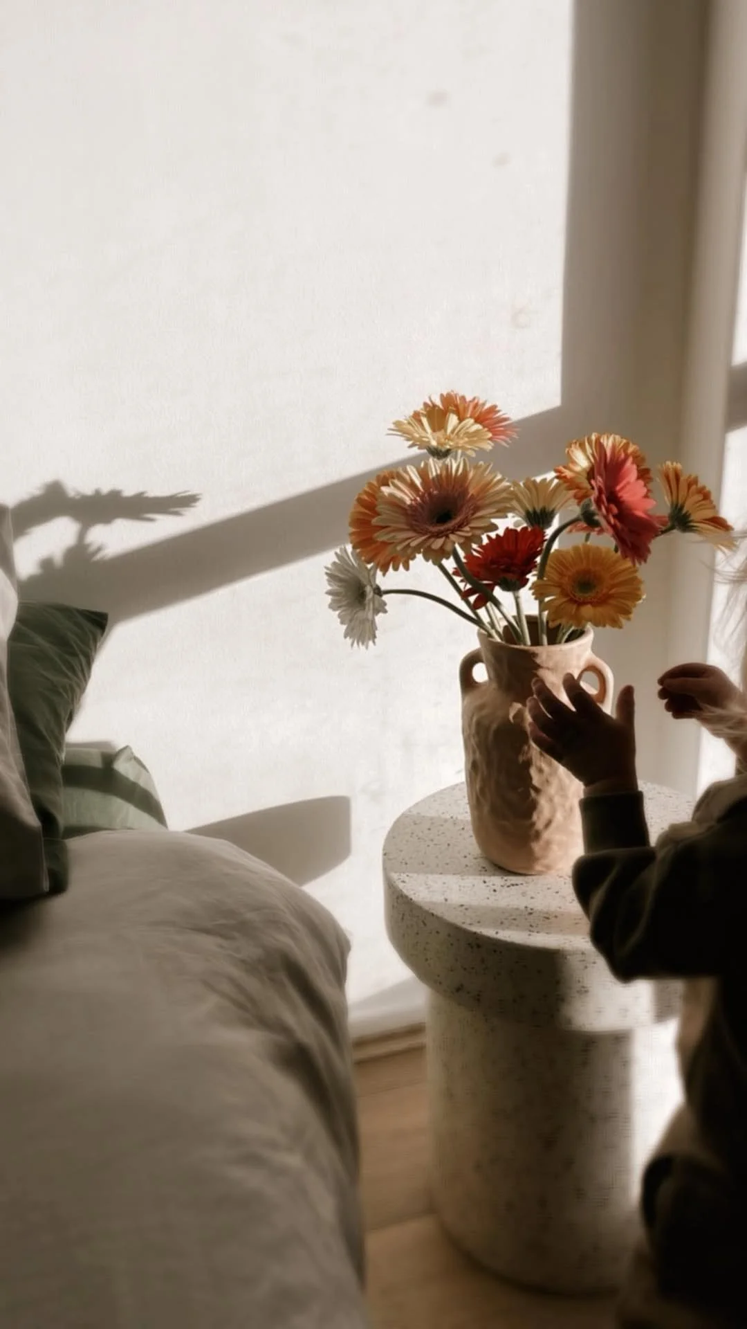 A person adjusting a bouquet of orange, yellow, and white flowers in a beige ceramic vase on a textured round side table, with shadows cast on a white wall from sunlight.