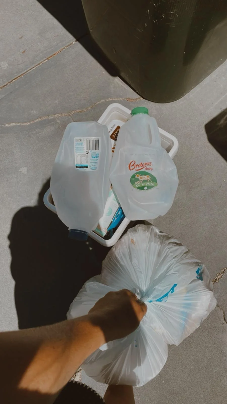A person holding a plastic bag filled with trash, with two empty plastic bottles on a white container and a trash can nearby.