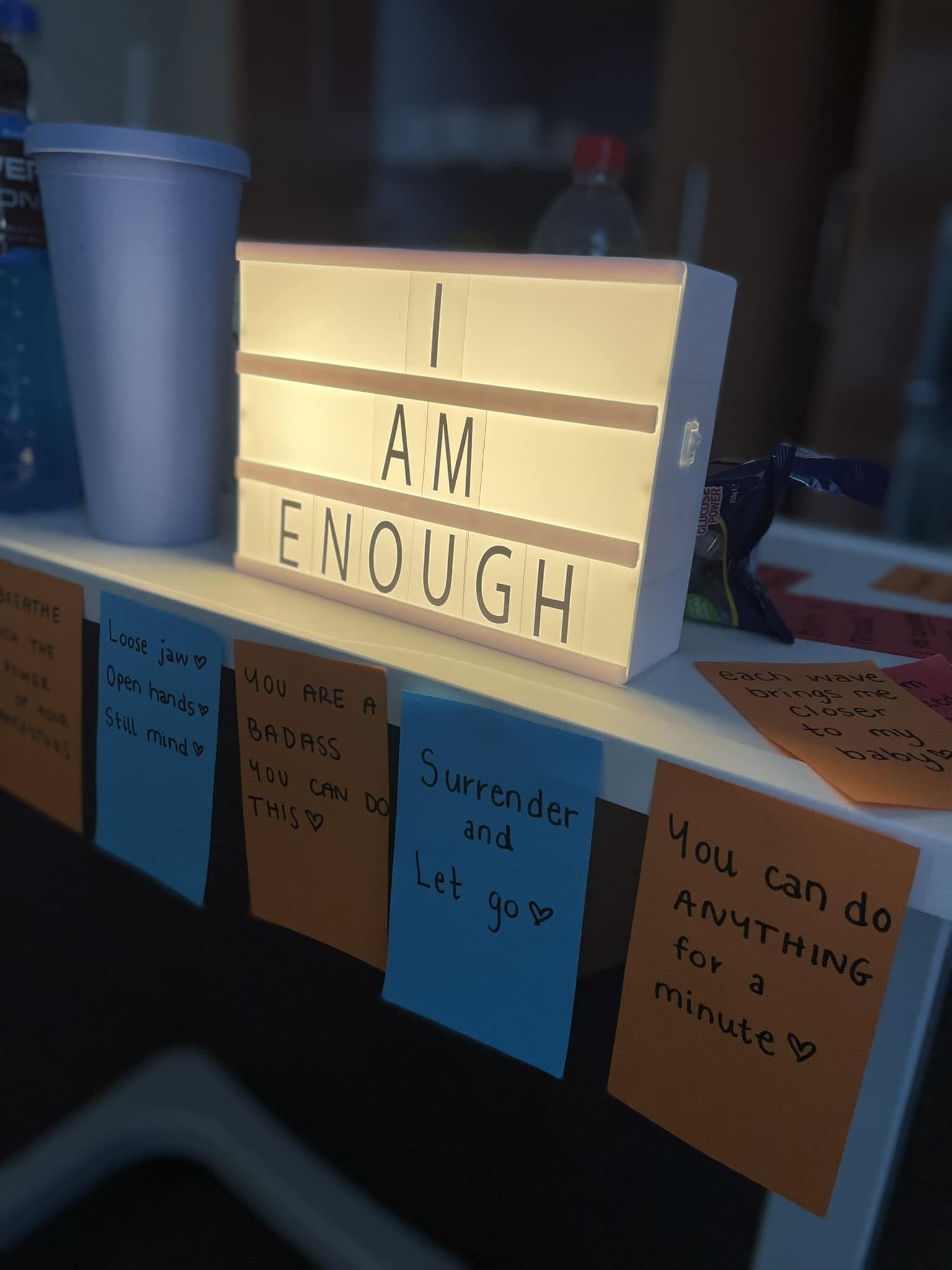 A lightbox sign displaying the message "I AM ENOUGH" on a shelf surrounded by colorful notes with motivational quotes.