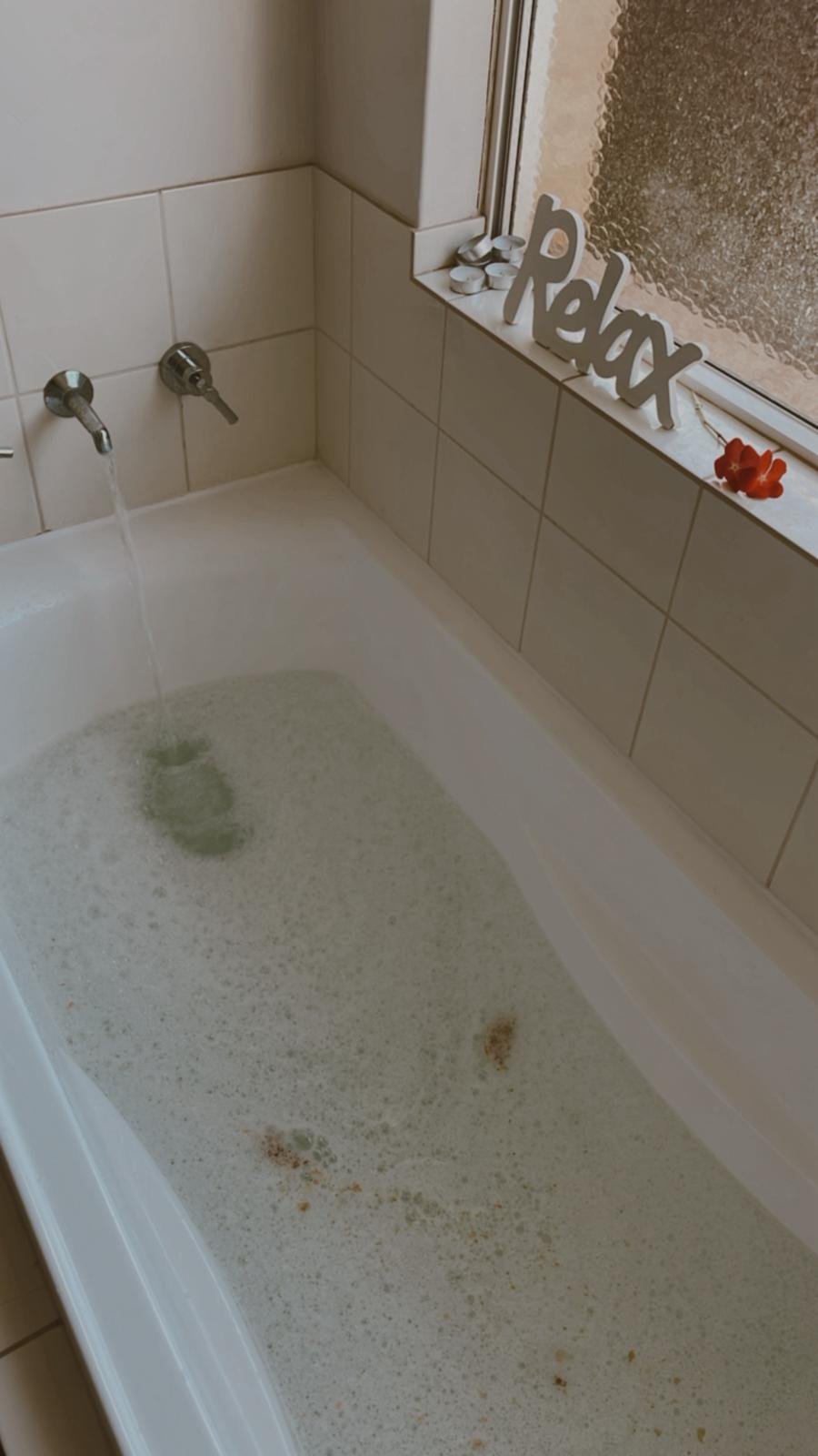 A bathtub filled with dirty, unclean water. There are two faucets, one flowing water into the tub. On the window sill are candles, a decorative 'Relax' sign, and some flowers.