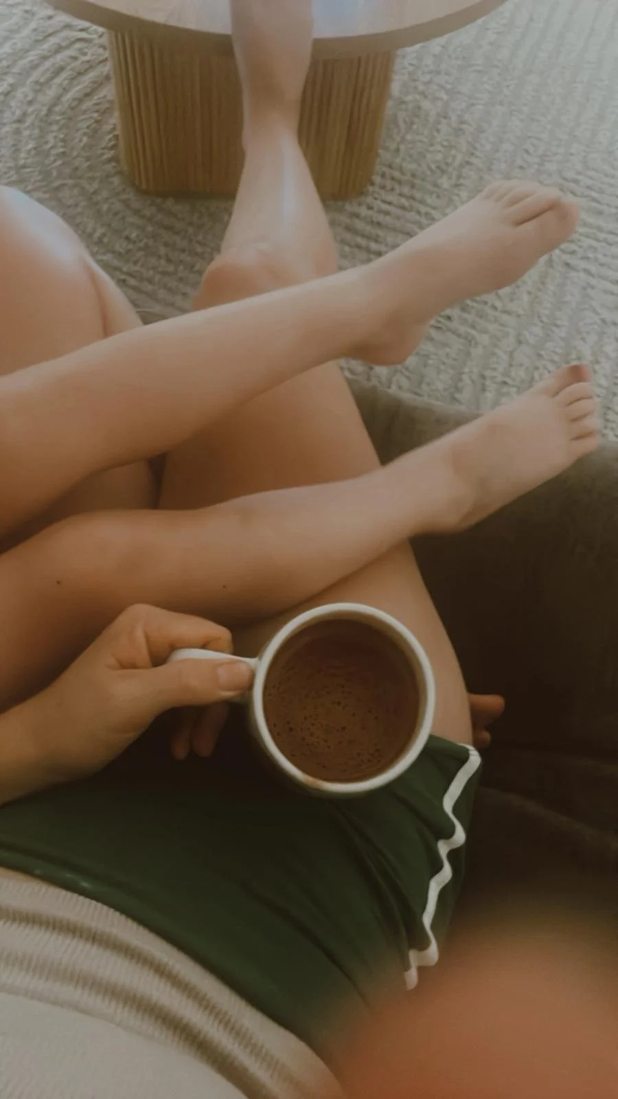 Person sitting on a sofa with legs crossed, holding a cup of coffee, with a small wooden table nearby and a carpeted floor.