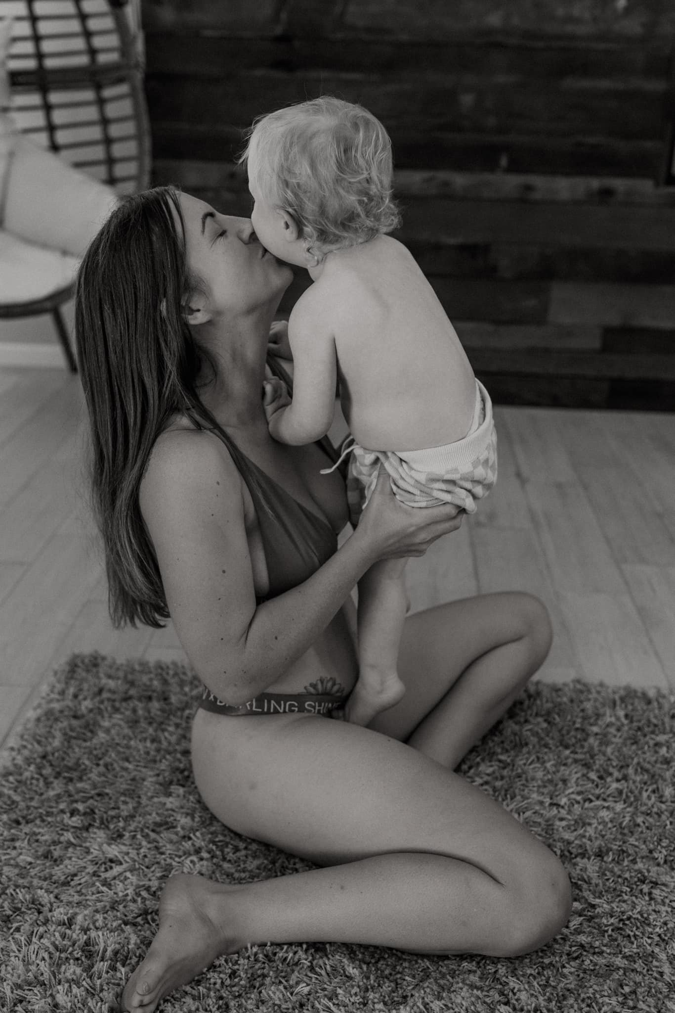 A woman with long hair kneels on a rug, lifting a shirtless young boy with curly hair, holding him close, as they kiss.