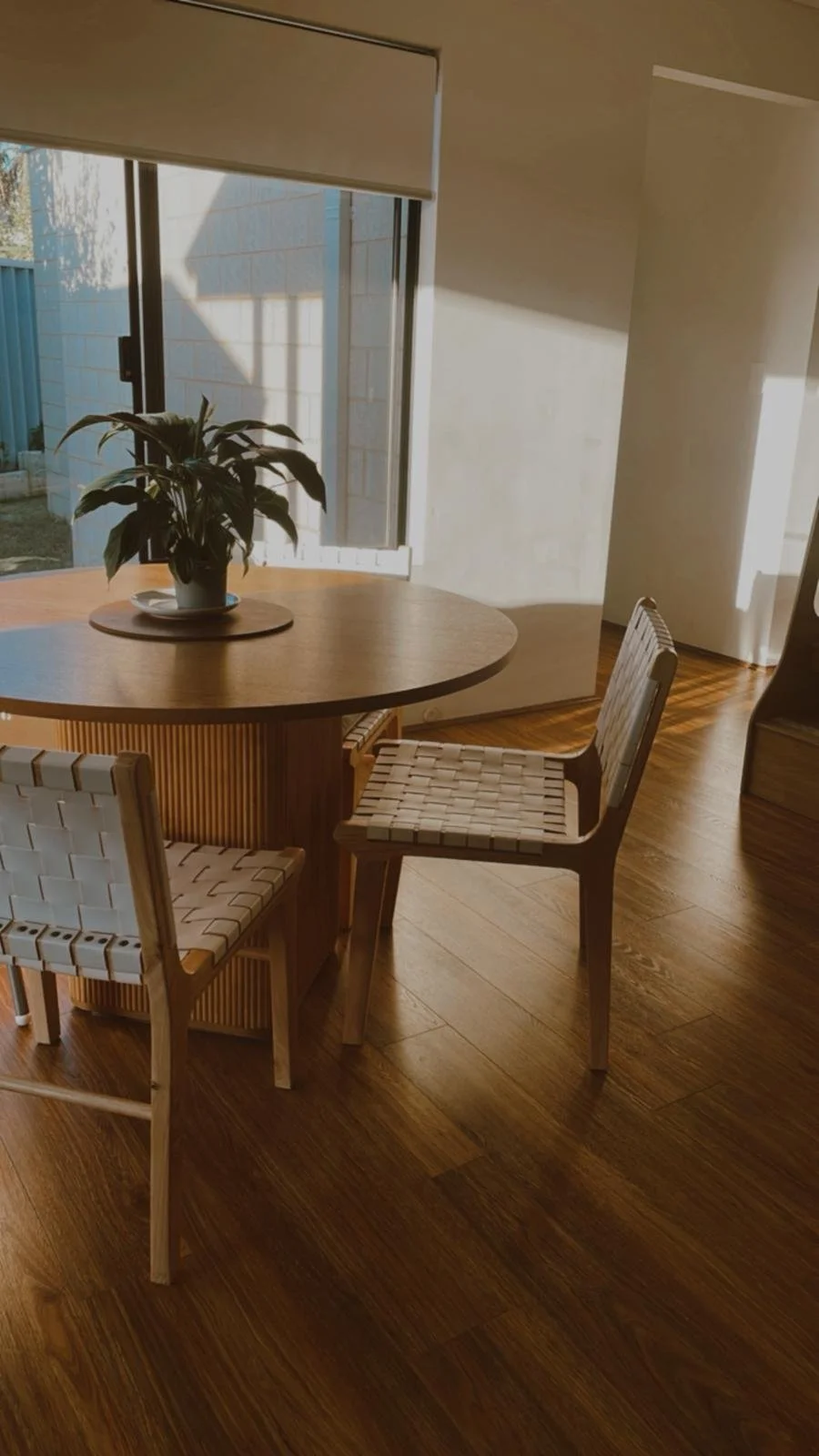 Wooden dining table with a potted plant and a plate on it, near a large window with a partially rolled-up blind, and hardwood floors.
