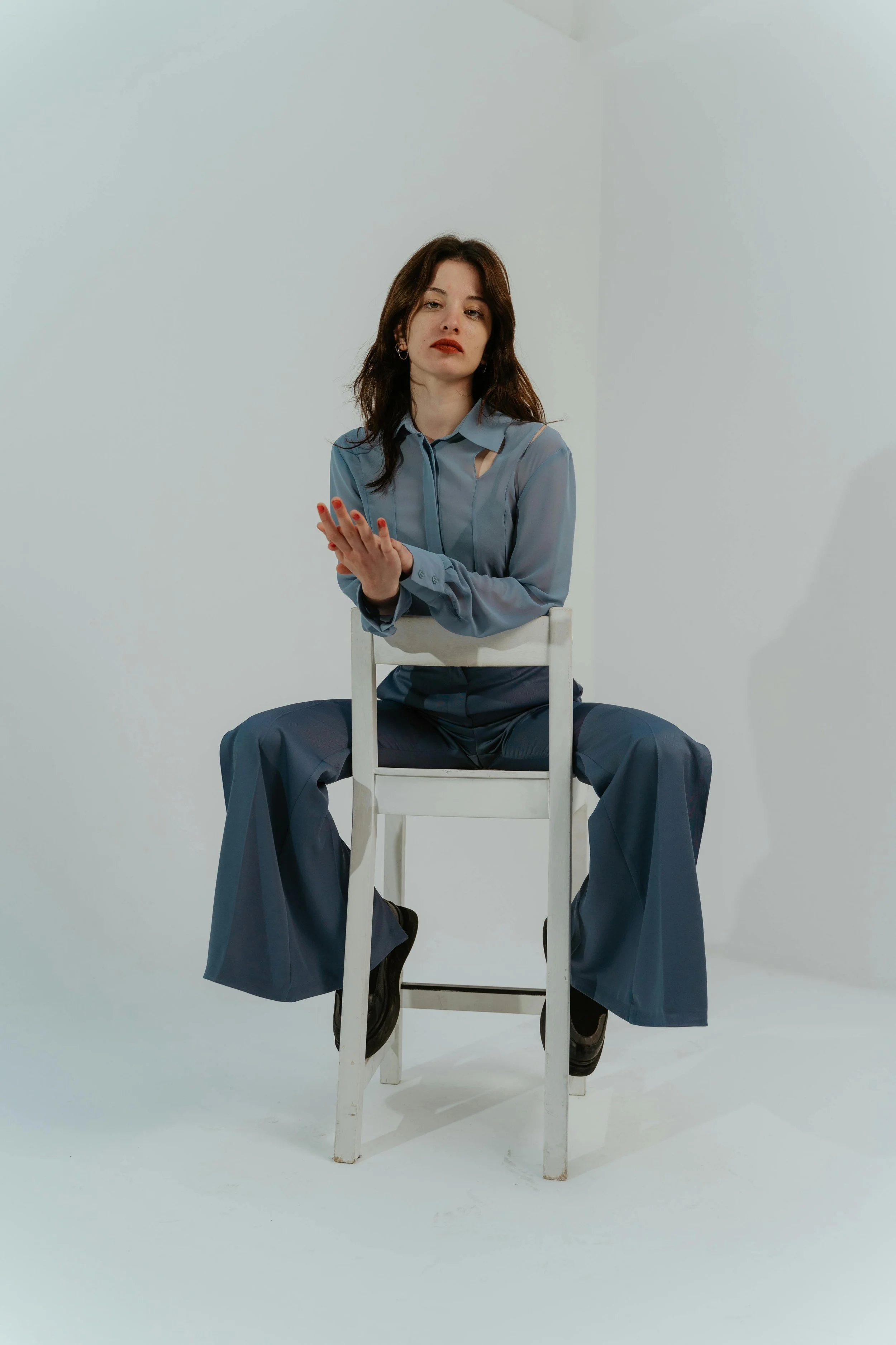 A woman sitting backwards on a white chair in a minimalistic setting, wearing a blue shirt and wide-leg blue pants, with dark brown hair and a neutral expression.