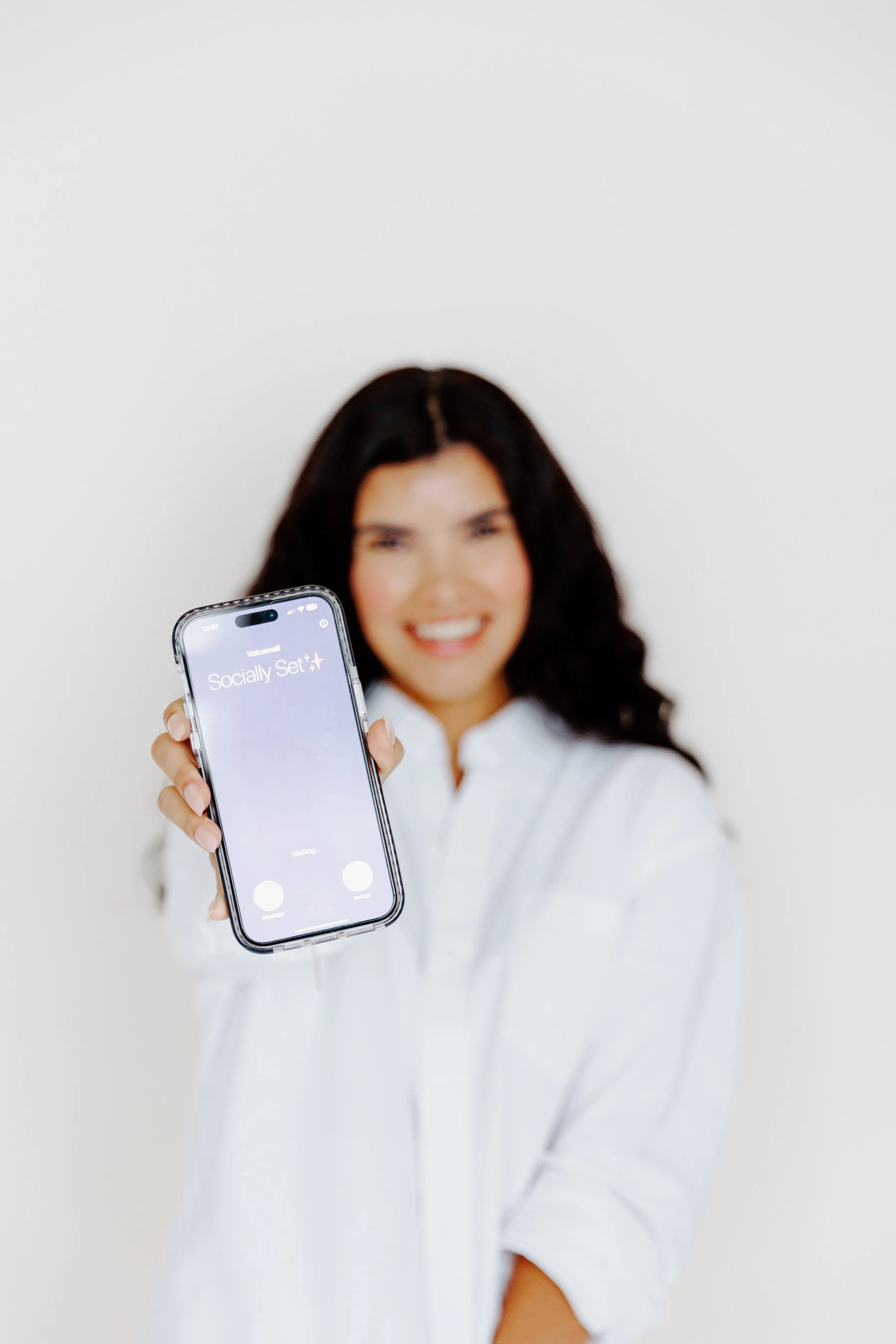 A woman with dark curly hair wearing a white shirt, smiling and holding up a smartphone showing an app screen with the text 'Socially Set'.