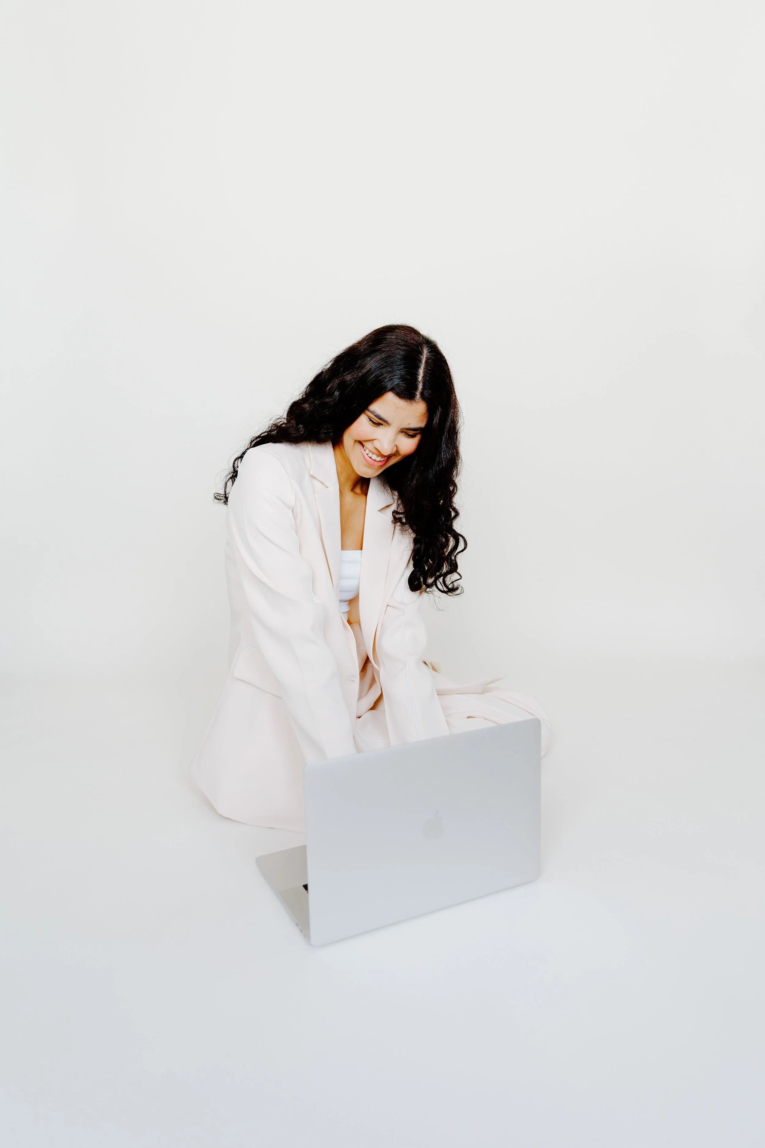 A woman with long black curly hair, wearing a white blazer, sitting on the floor and smiling at a silver laptop in front of her against a plain white background.