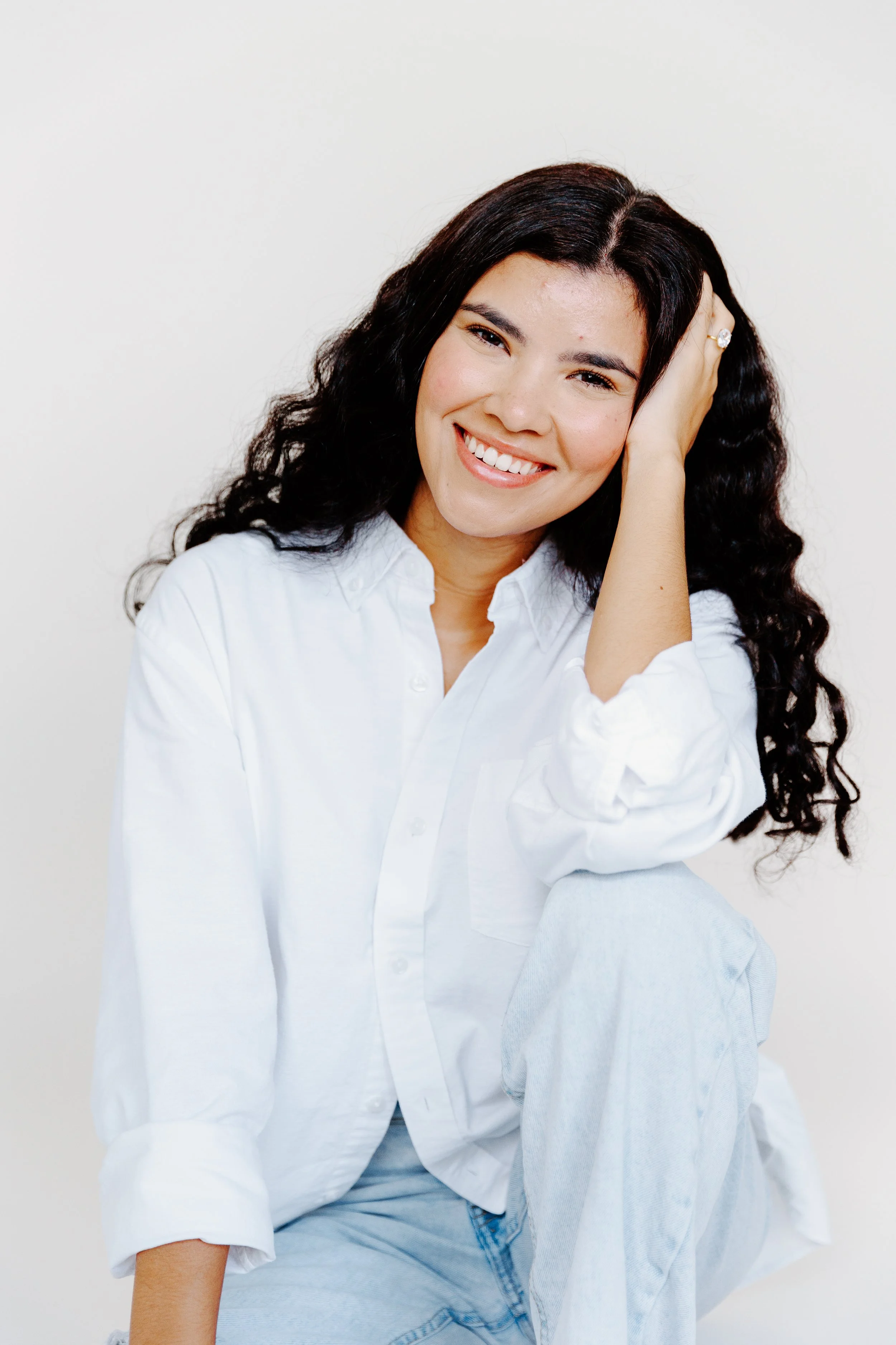 Smile woman with black, curly hair, wearing a white shirt and light blue jeans, sitting against a plain white background.