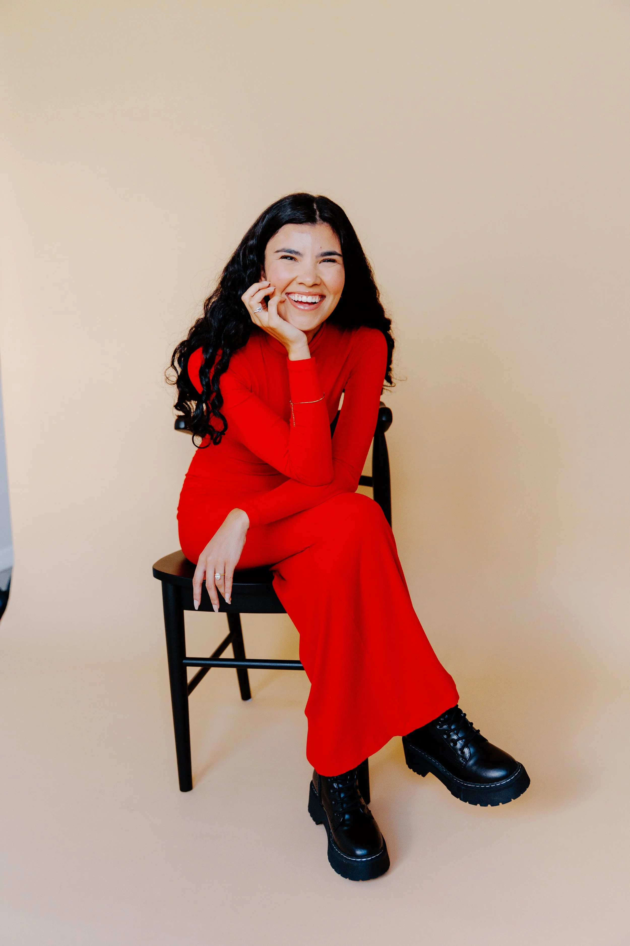 A woman with long curly black hair wearing a red dress and black chunky boots, sitting on a black chair, smiling and resting her face on her hand against a beige background.