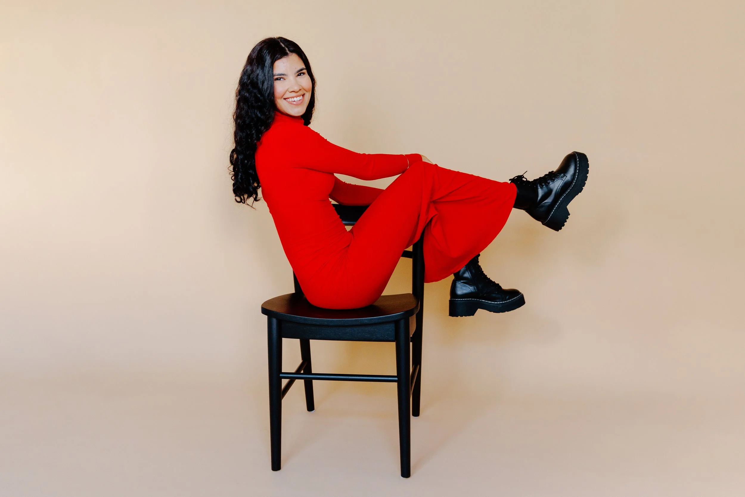 Woman sitting sideways on a black wooden chair against a beige background, dressed in a red outfit and black lace-up boots, smiling at the camera.