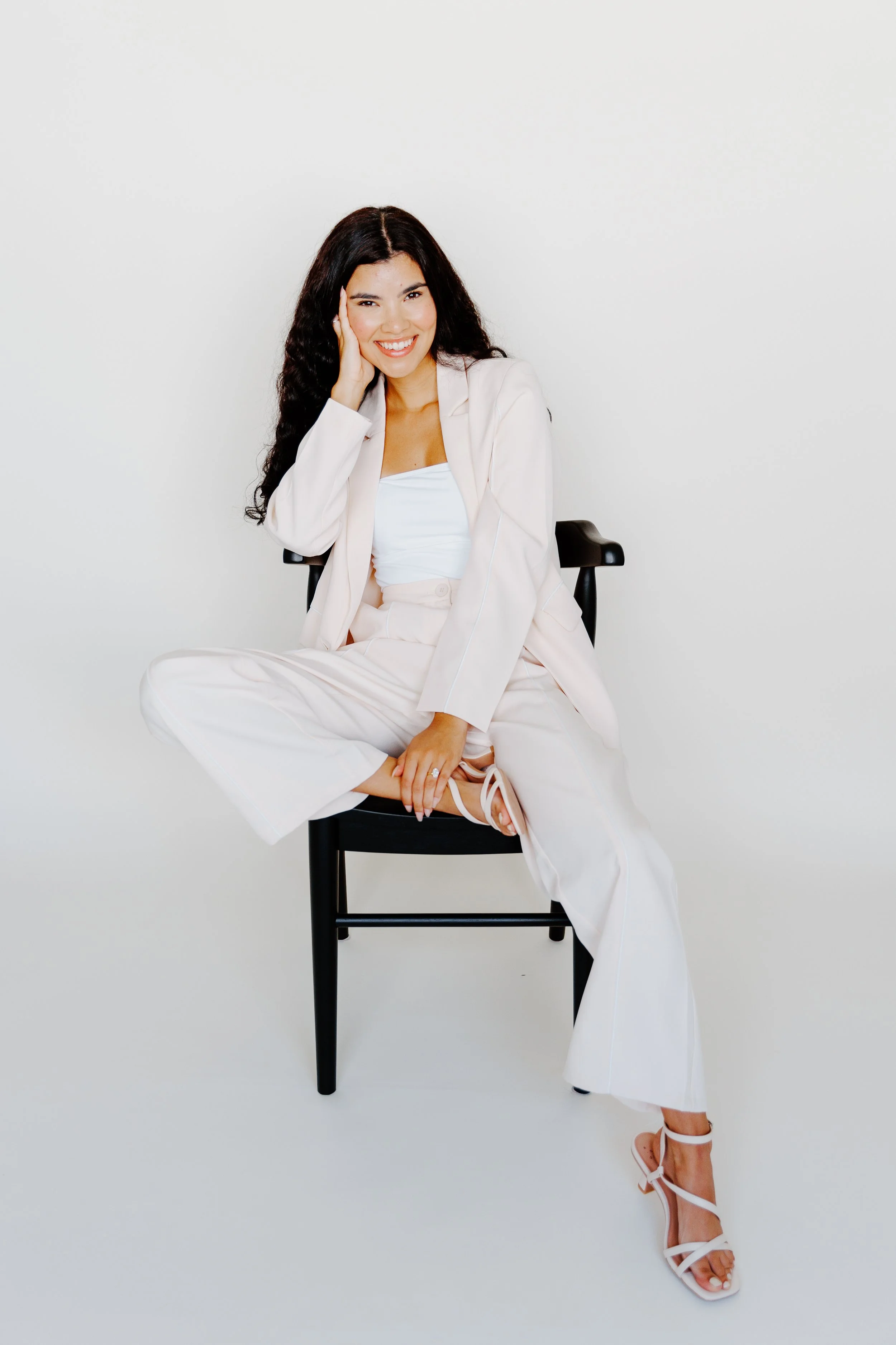 A woman with long dark hair sitting on a black chair against a plain white background, smiling and dressed in light-colored formal attire with high-heeled sandals.