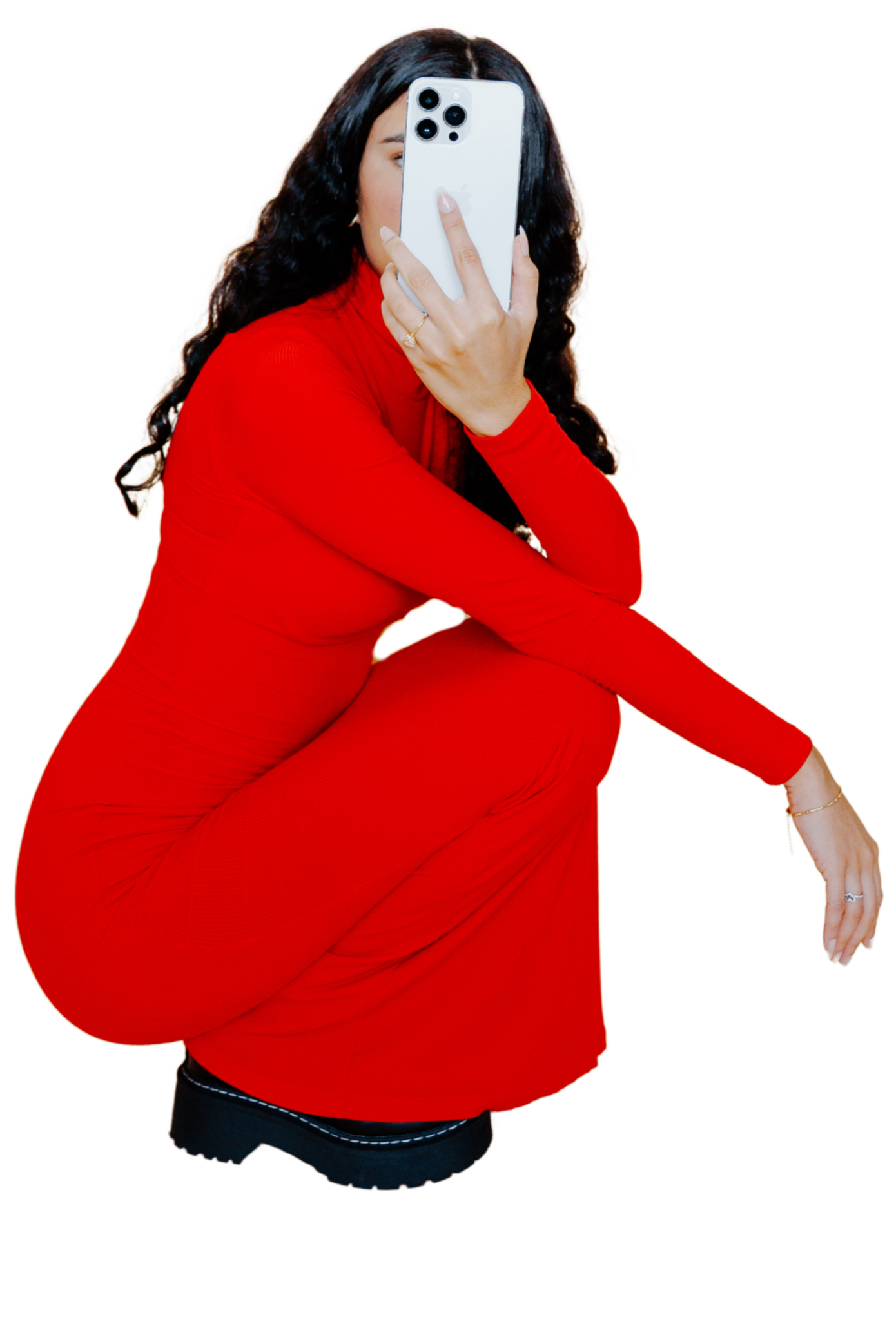 A woman crouching in a red outfit taking a selfie with a white iPhone, with long wavy dark hair and accessories including rings and bracelets.