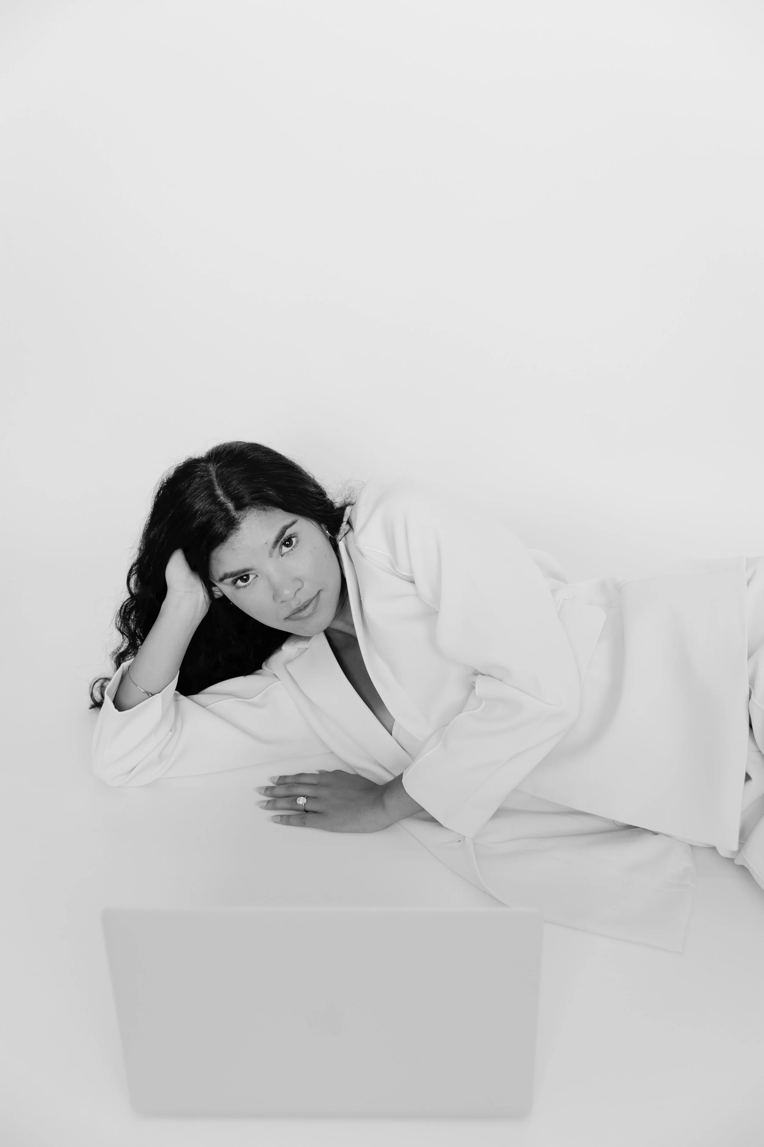 Black and white photo of a woman lying on her side, looking at the camera, with a laptop in front of her, against a plain background.