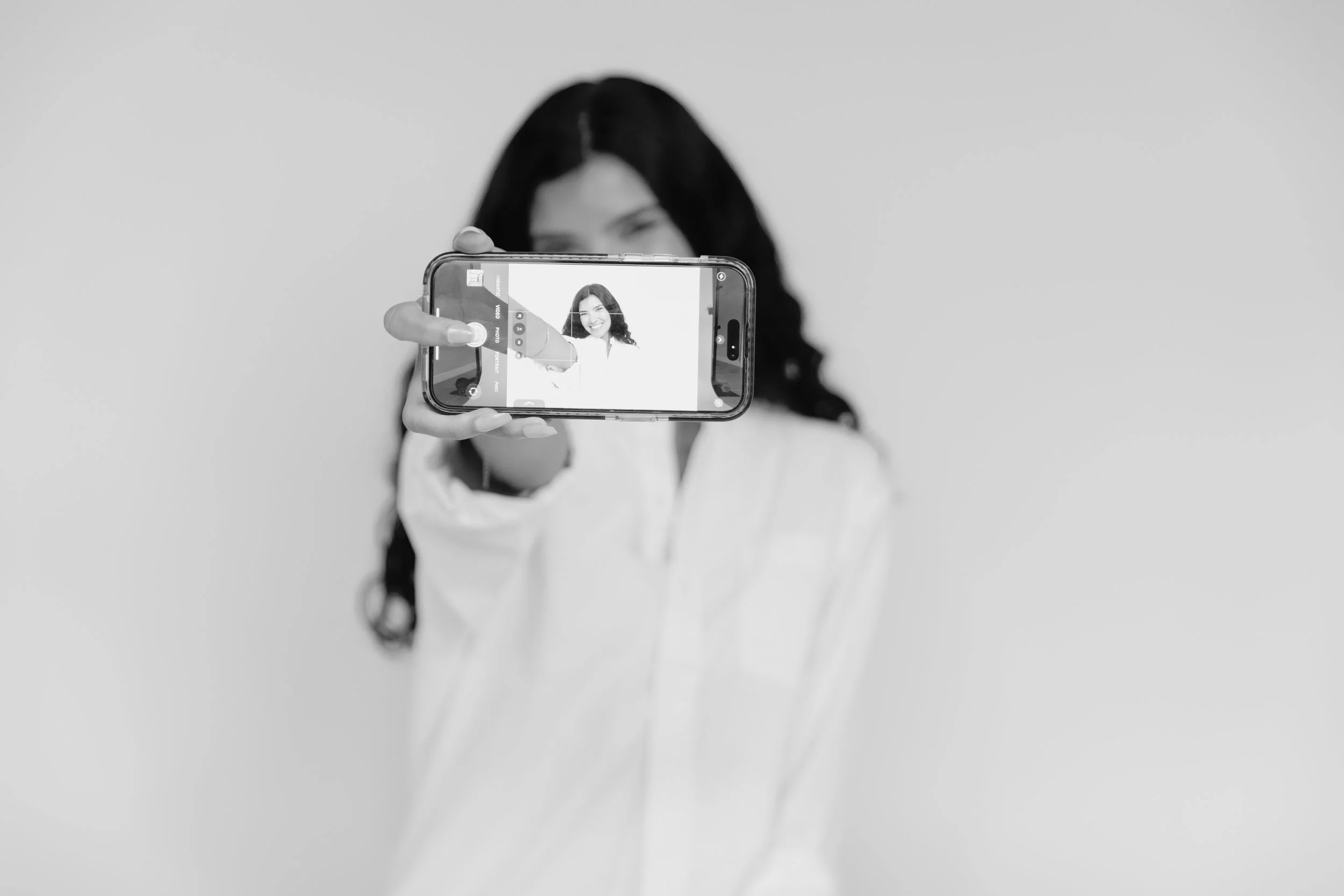 A woman with dark hair taking a selfie with a smartphone against a plain background.