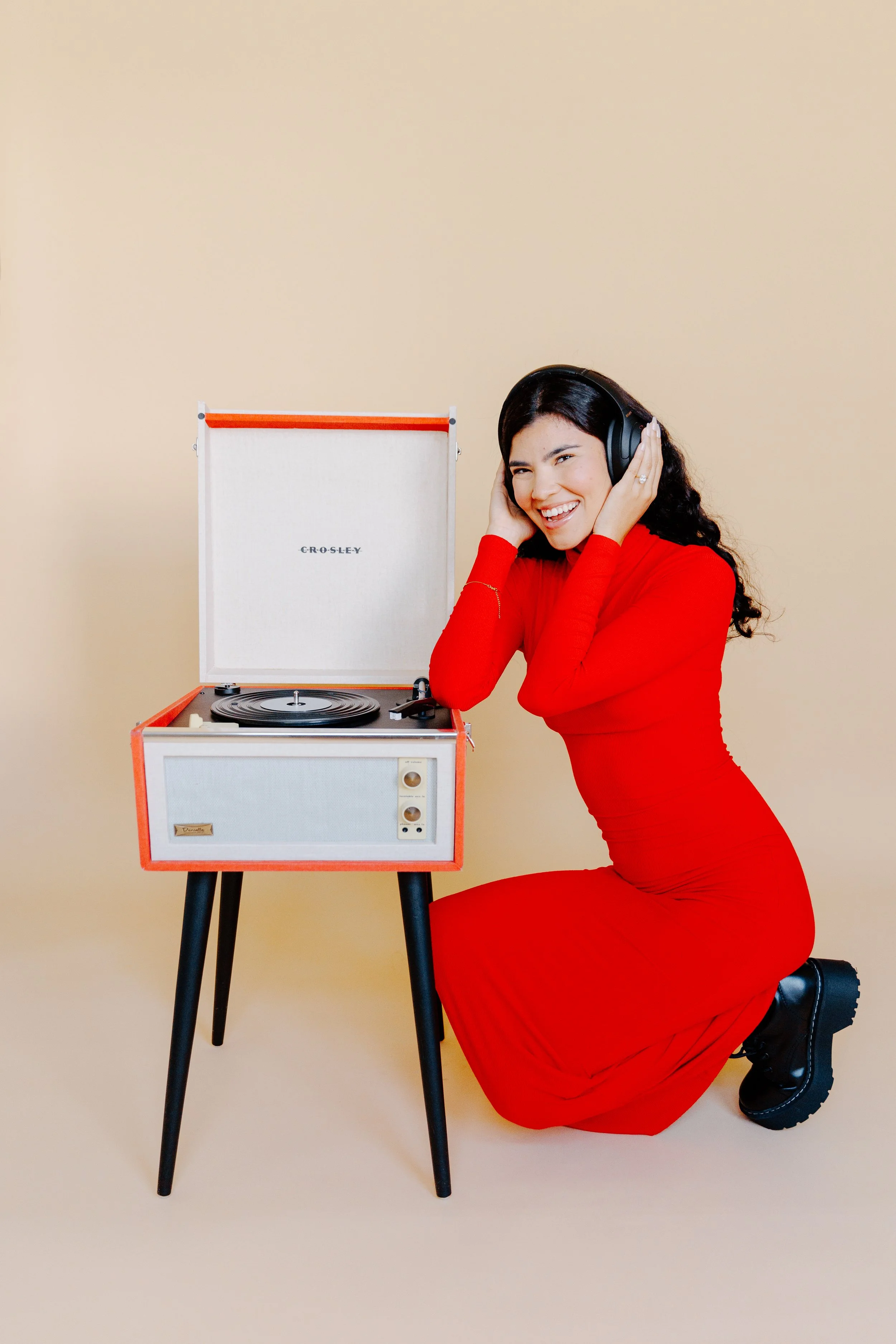 A woman in a red dress kneeling on the floor, listening to music through headphones, next to a vintage portable record player against a beige background.