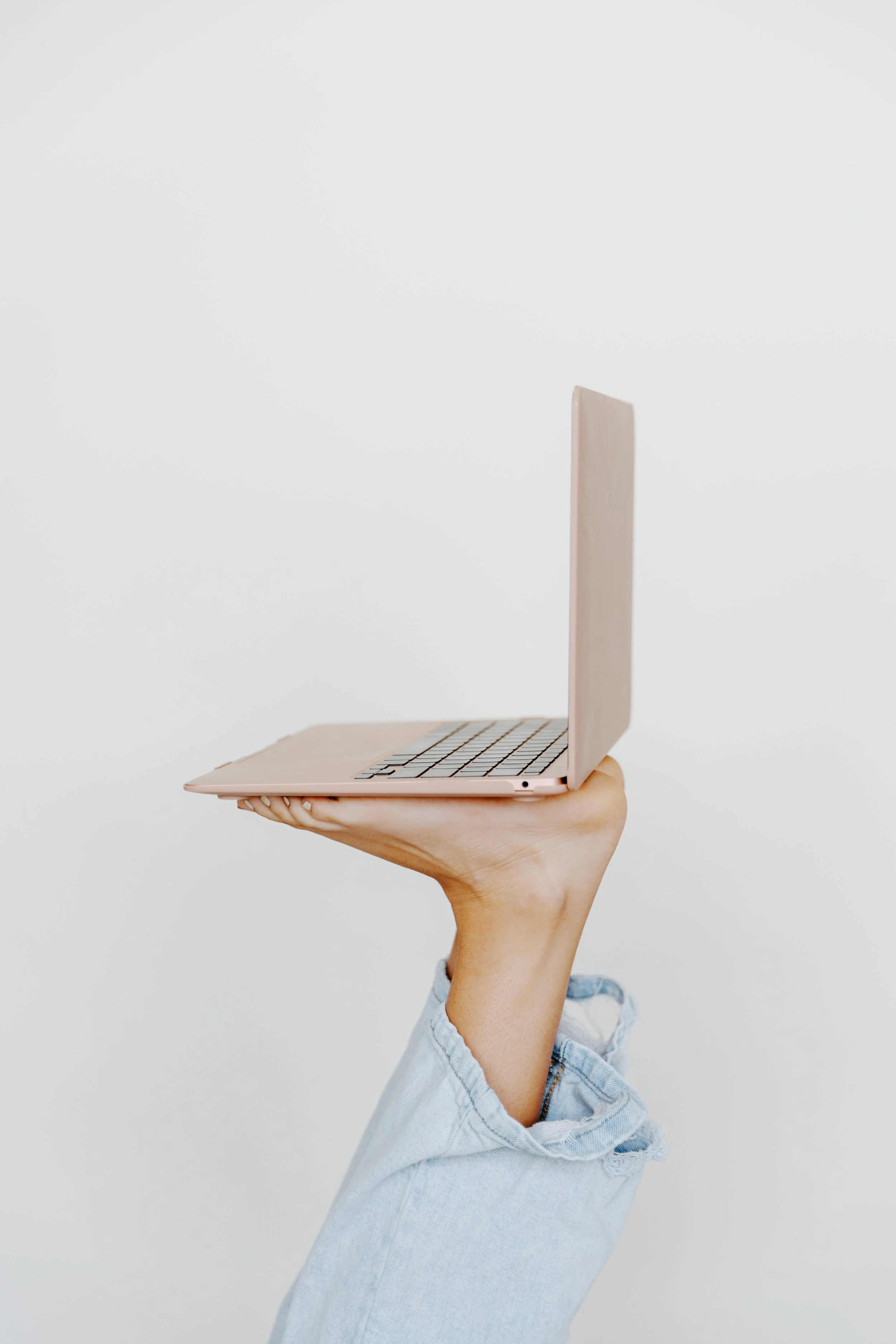A person holding a pink laptop in one hand against a plain white wall.