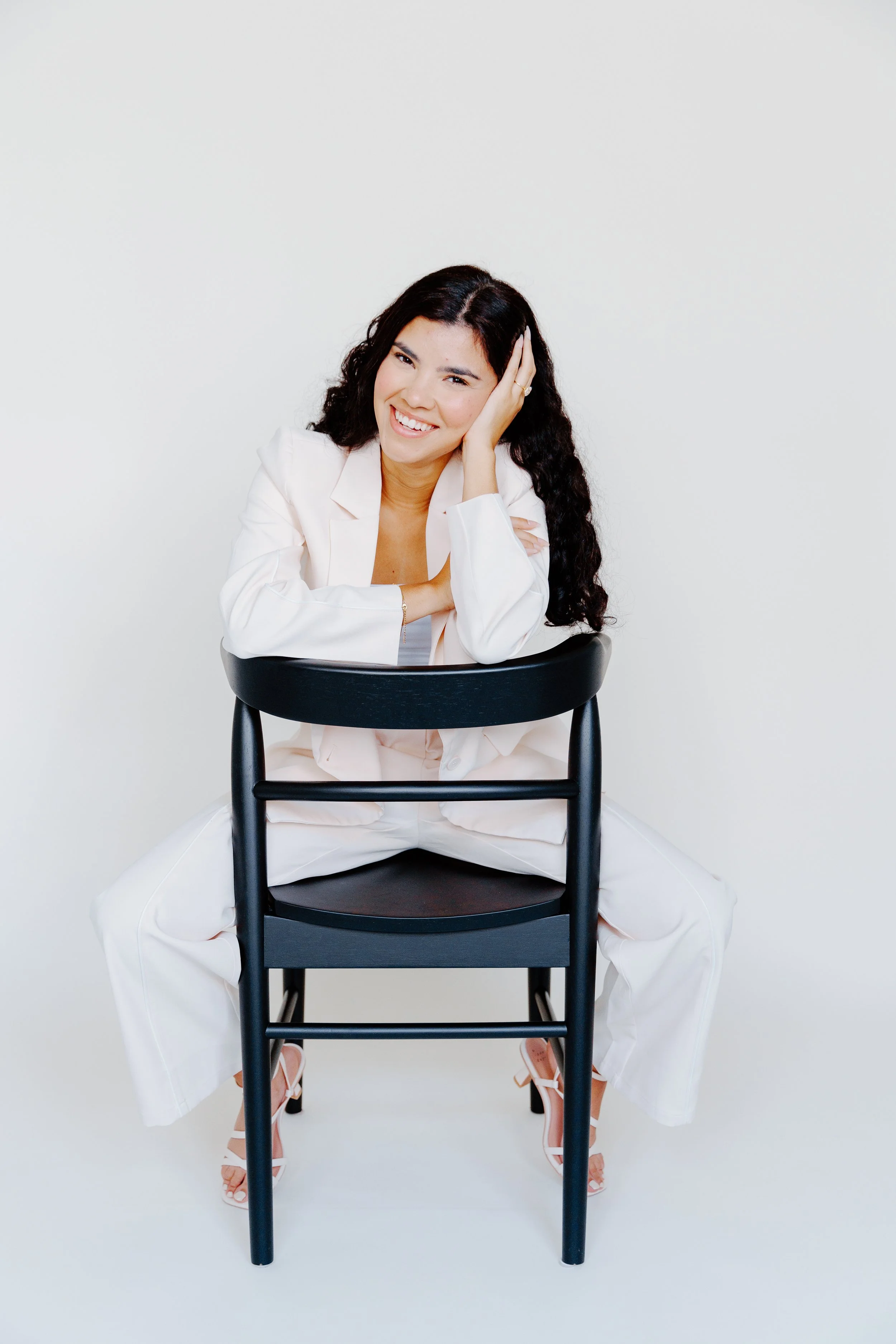 A woman with long curly black hair, wearing a light pink blazer and pants, is sitting on a black chair with her legs open. She is smiling and resting her head on her left hand, with her right arm on the middle of the chair.