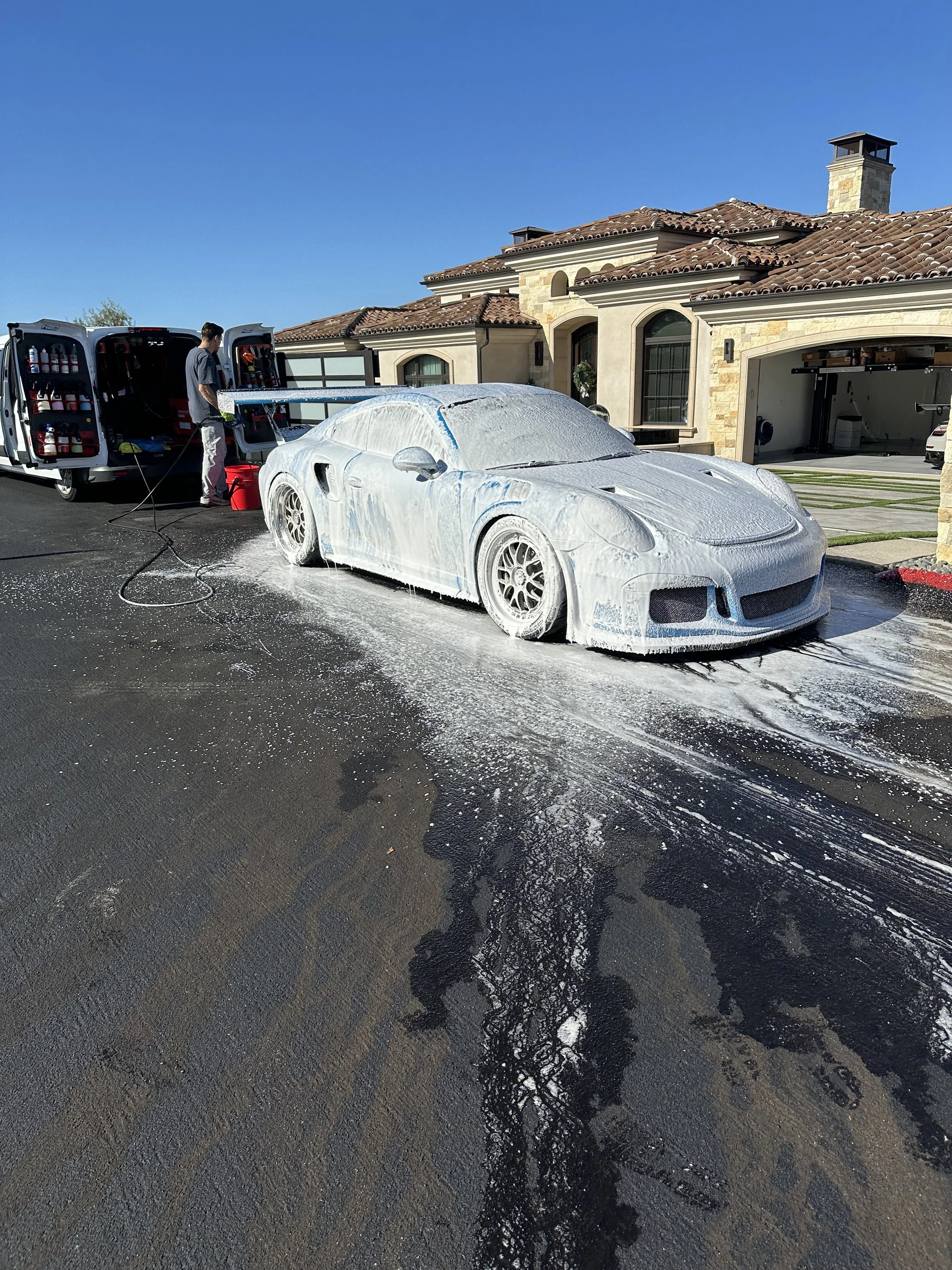 A white sports car covered in foam being washed in the driveway of a large house with a red-tiled roof and stone exterior. A person is using a hose to spray water on the car, and two vans with cleaning supplies are parked nearby.