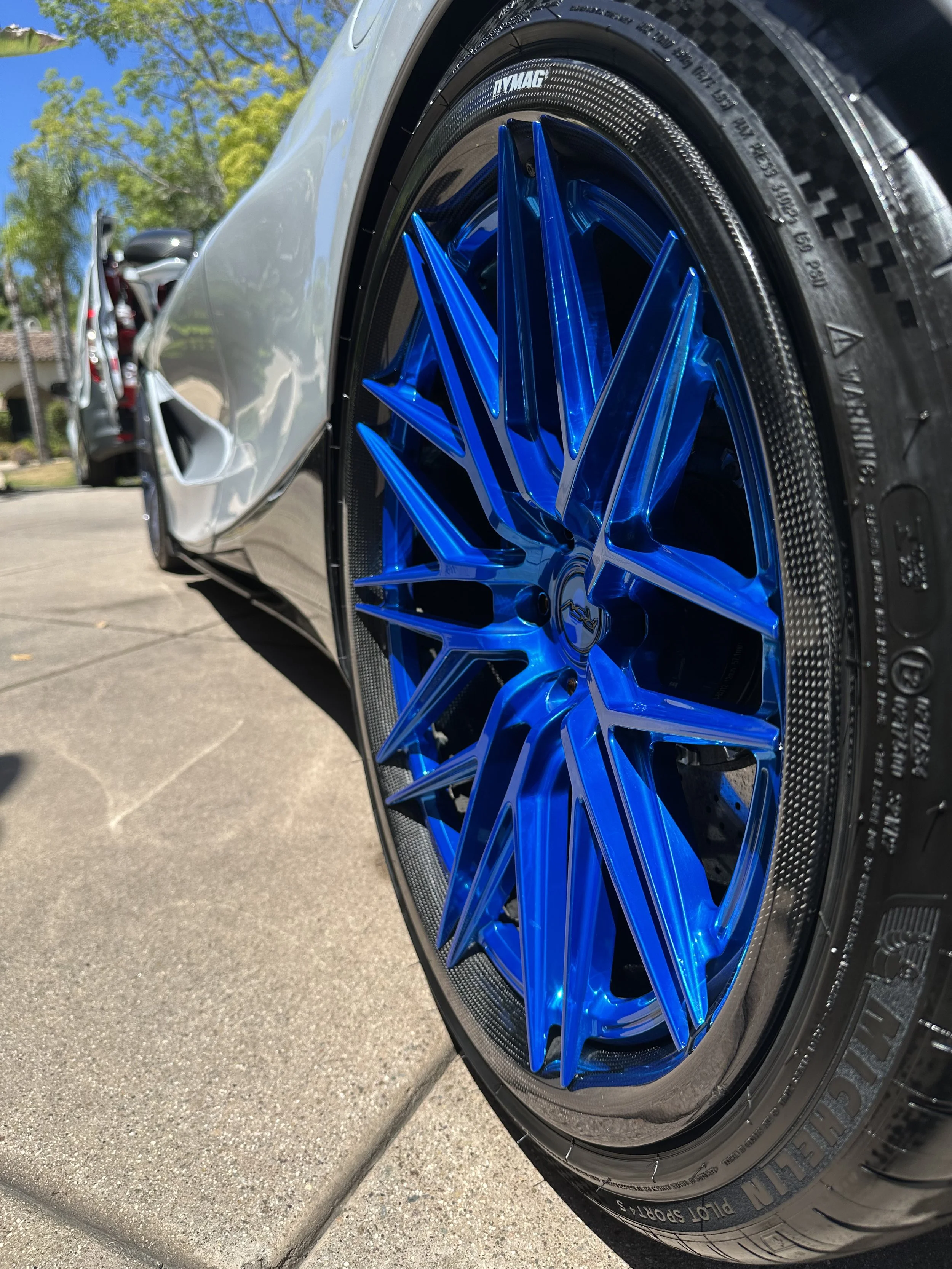 Close-up of a sports car's bright blue custom wheel and tire, with part of the silver body and a car with an open hood in the background under clear skies.