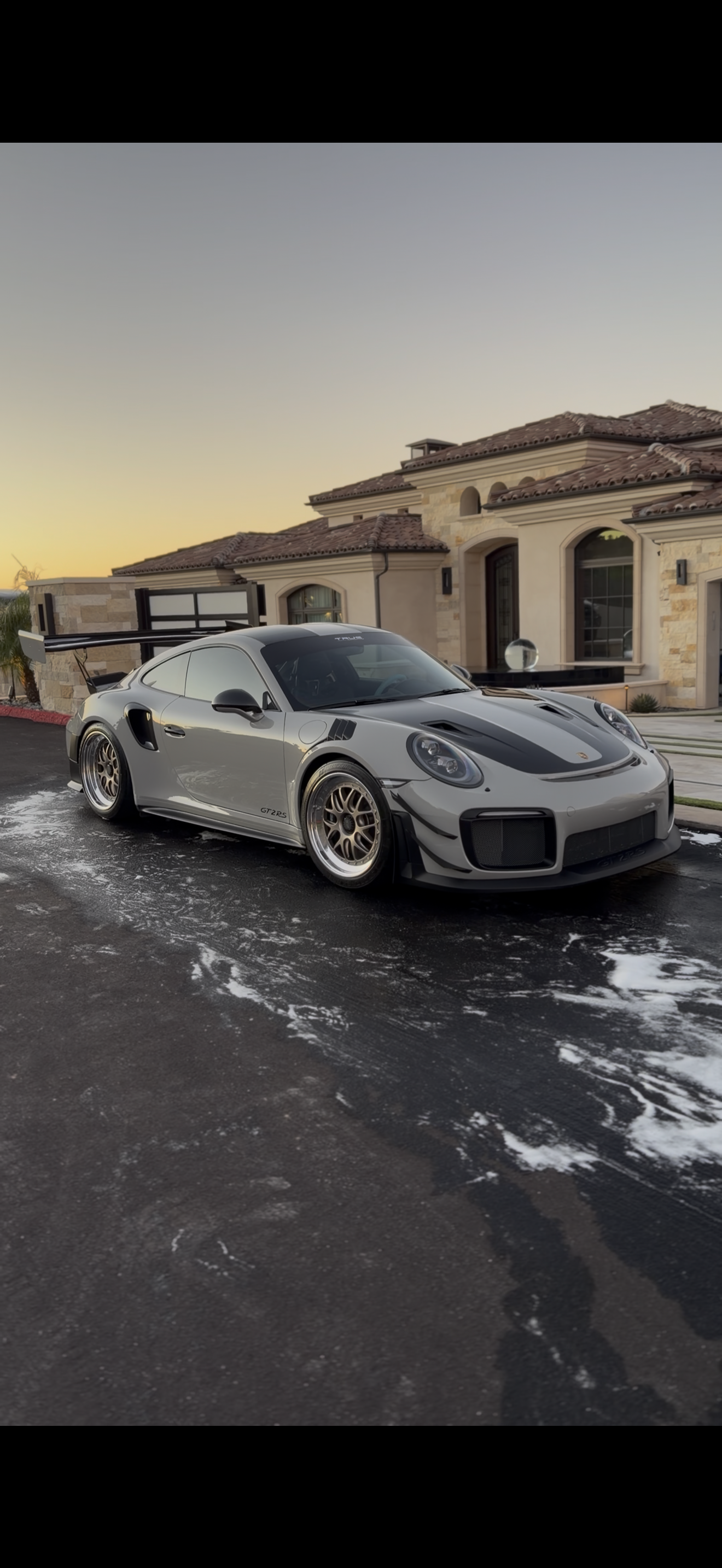 A silver Porsche 911 GT2 RS with black accents, aftermarket wheels, and a large rear wing parked on a driveway with soap and water, house with stone accents and red tile roof in the background at sunset.