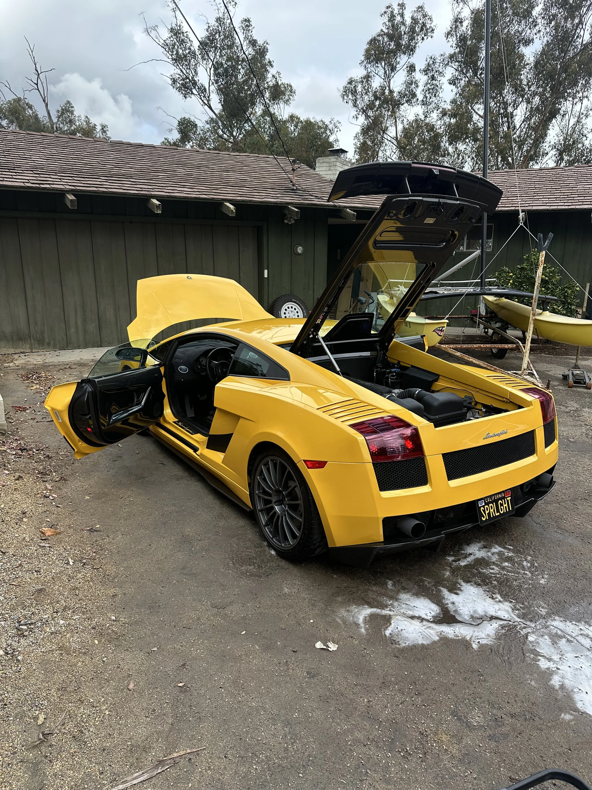 Yellow Lamborghini sports car with open driver door and rear hood open, parked outside on dirt ground near a green building and trees.