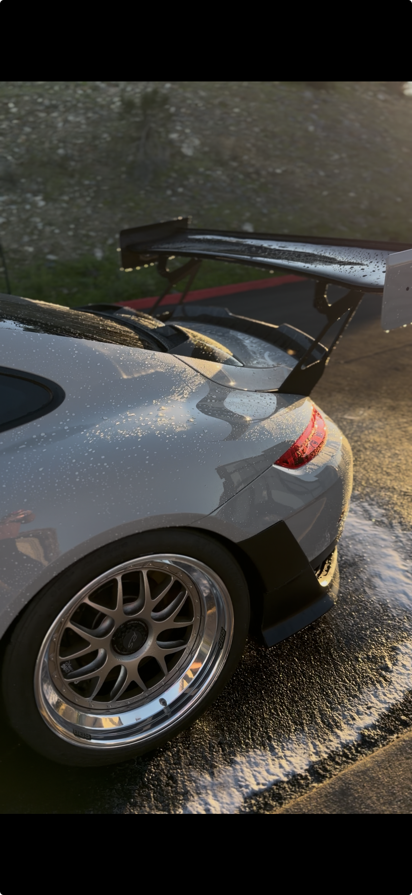 Close-up of a Porsche GT2 RS with a large rear wing, water droplets on the surface, and a wheel with a multi-spoke design, parked on top of a hill in beautiful Rancho Santa Fe.