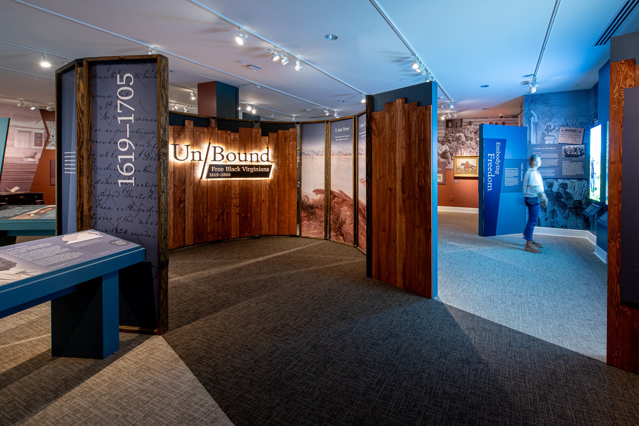 Exhibit in a museum with wooden panels and blue display stands, one labeled 'Un/Bound' about black Virginians 1619-1865, a visitor looking at a display, and informational panels about freedom and history.