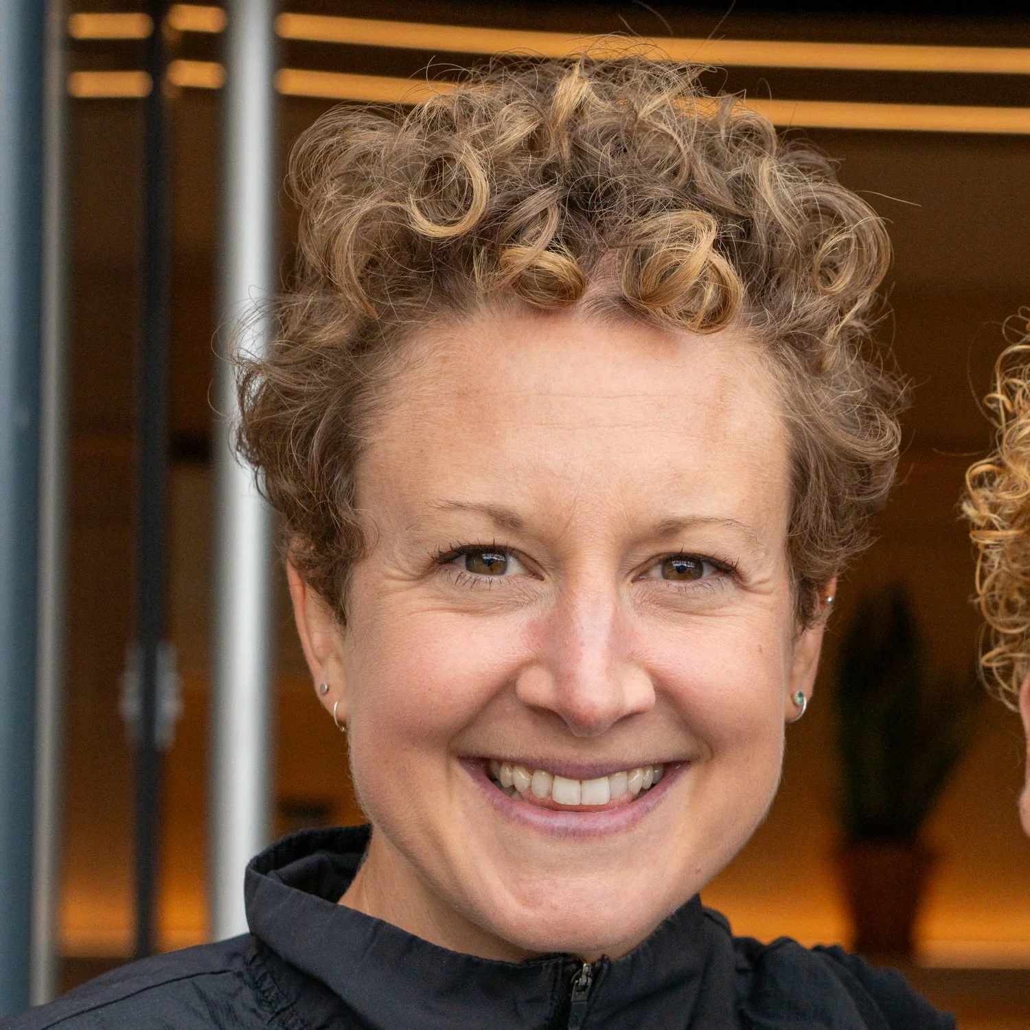Close-up photo of a smiling woman, Maria the NuMi founder. She has short curly light brown hair and is wearing a black jacket, and standing outside NuMi HQ with wooden shelves in the background.