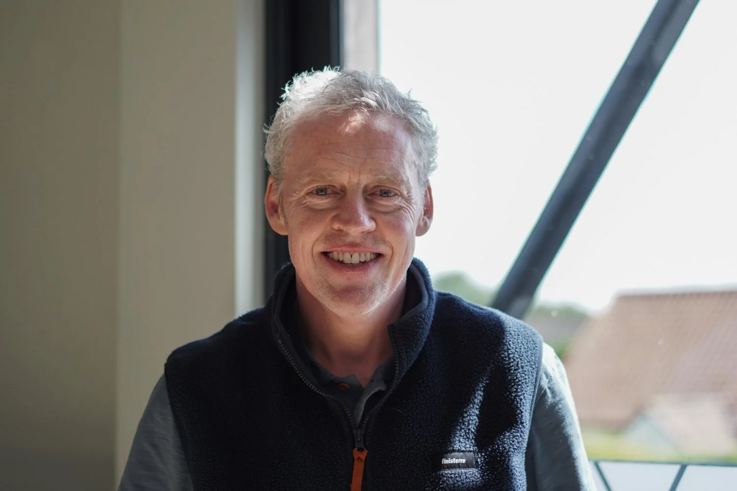 Smiling older man with gray curly hair sitting indoors near a window with a background of rooftops and sky.