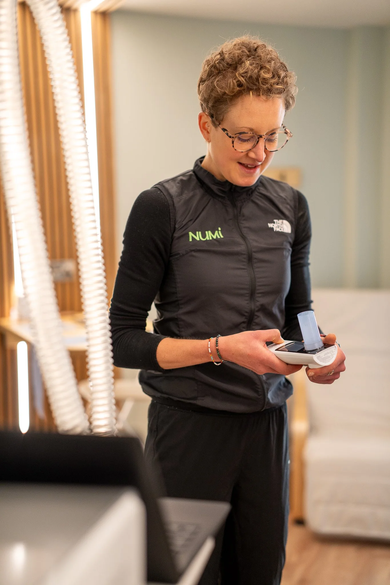 Close-up photo of a smiling woman, Maria the NuMi founder. She has short curly light brown hair and is wearing a black jacket, and standing outside NuMi HQ with wooden shelves in the background.
