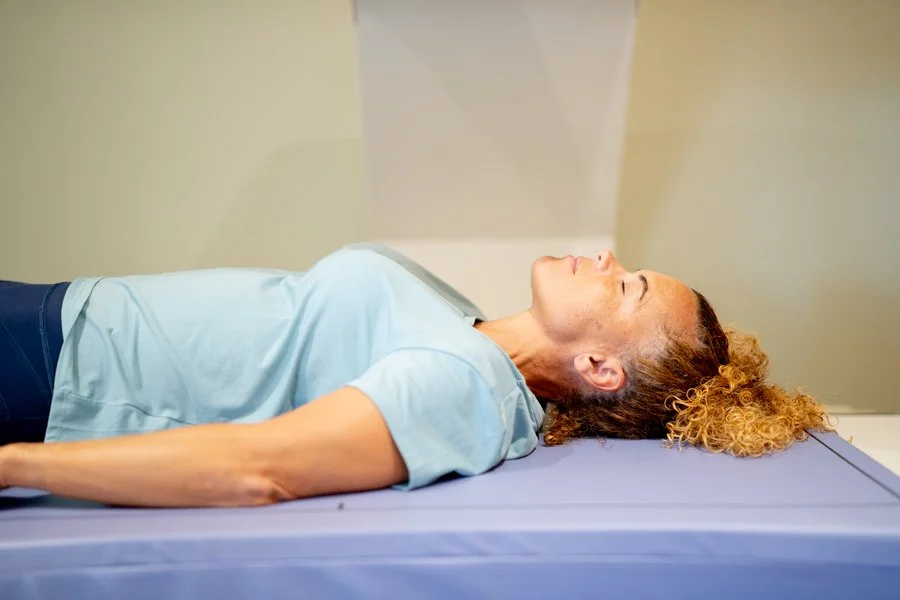 A woman lying on her back on a DEXA scanner, smiling and looking up at the ceiling, with a plain wall background.