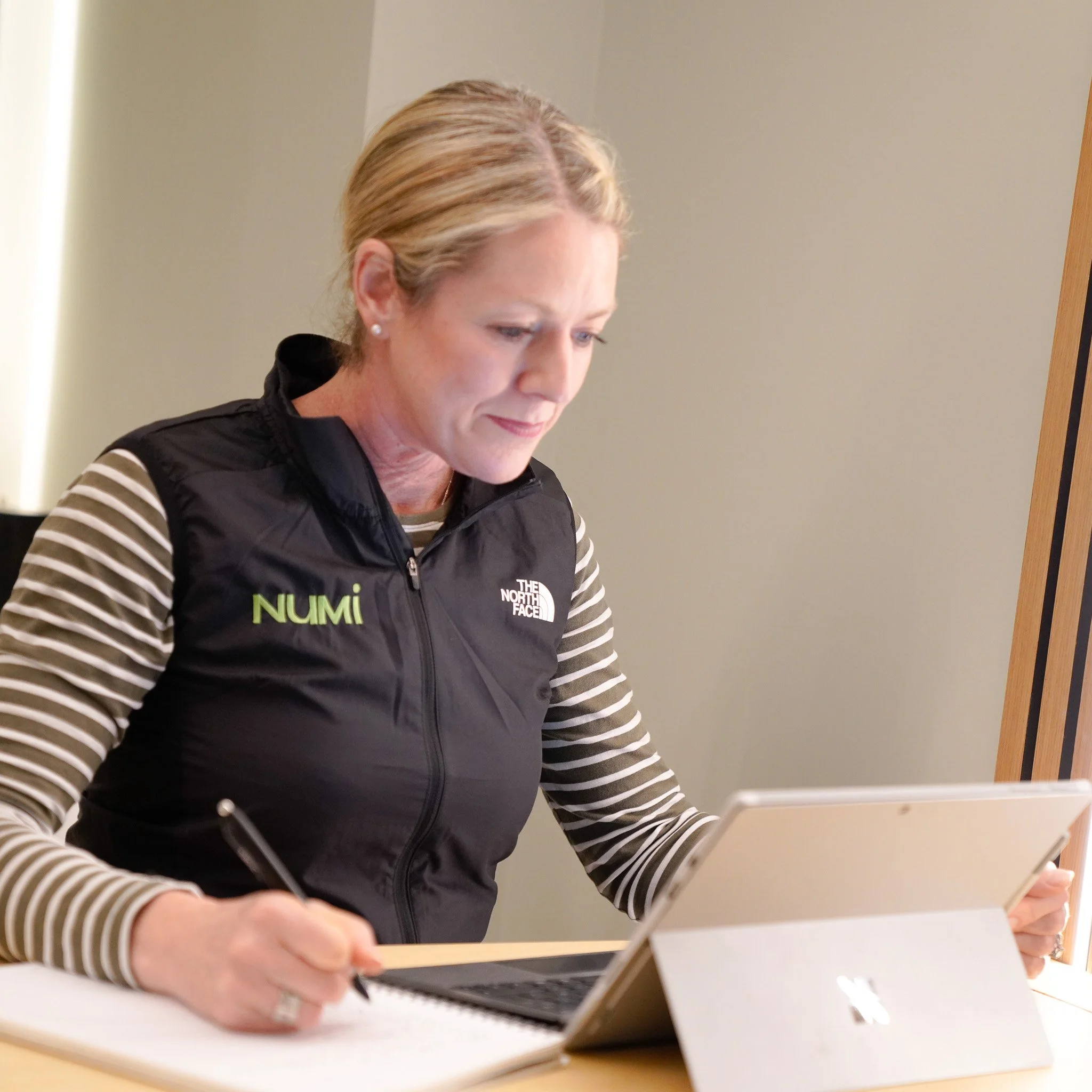 A woman working at a desk with a laptop and notepad, wearing a black North Face vest with 'NUMI' embroidered, and a striped long sleeve shirt.