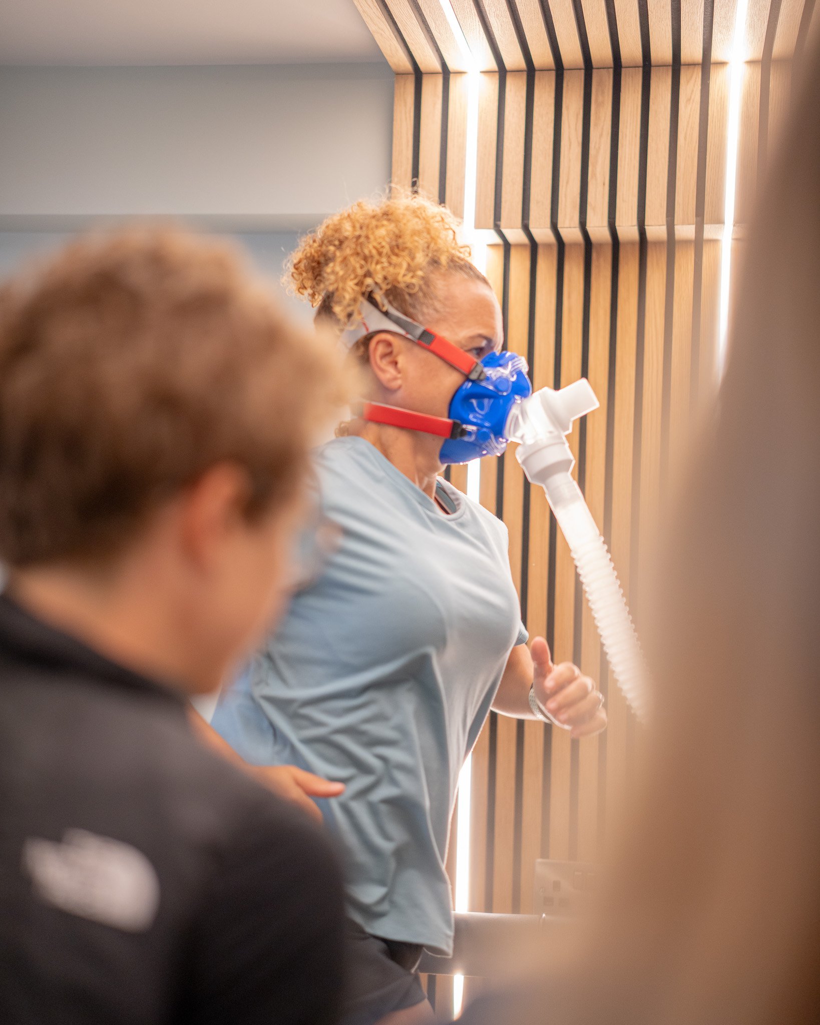 A woman with curly hair wearing a blue mask connected to a white tube, running indoors against a wooden slat wall, with another person partially visible in the foreground. She is performing a VO2 max and fatmax test.