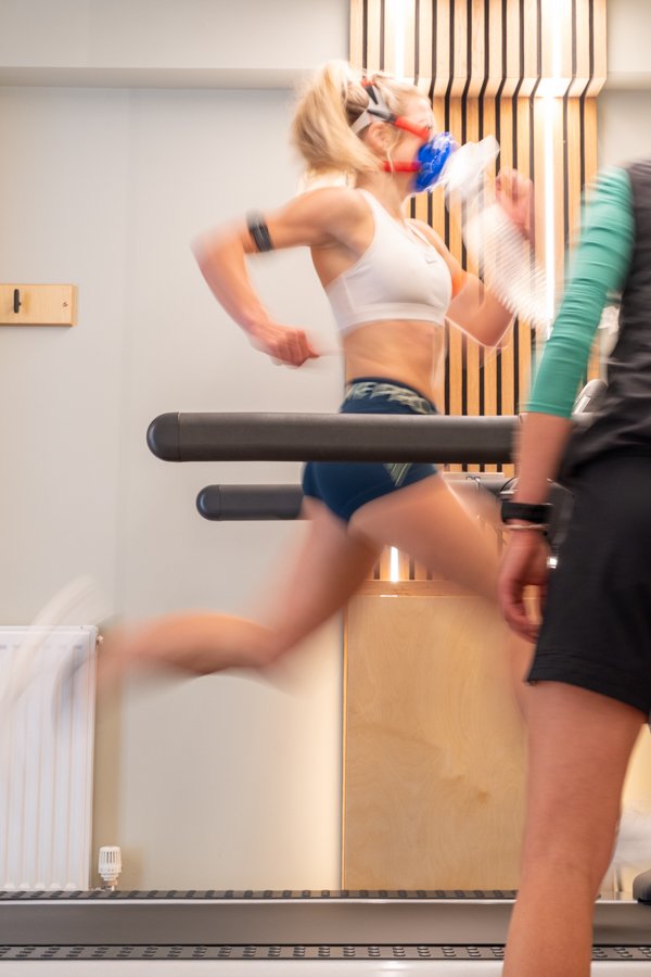 A woman running in a gym wearing a face mask and workout clothes, with a trainer nearby.