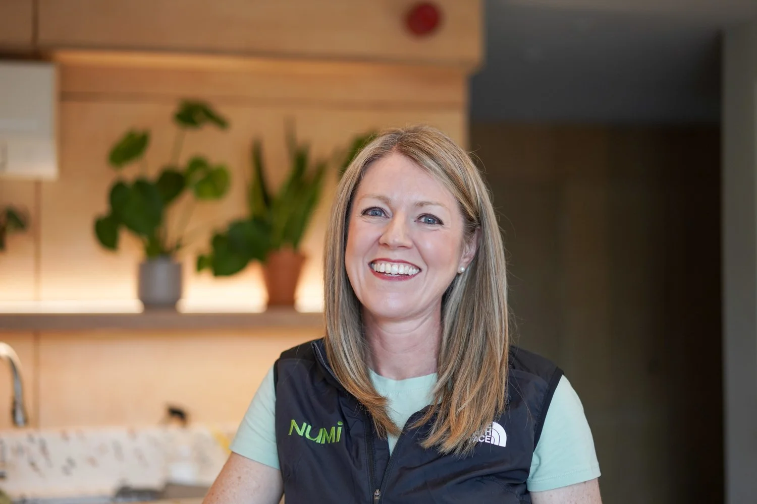 A smiling woman with shoulder-length blonde hair wearing a light green shirt and a black vest with 'NUMI' and 'The North Face' logos, standing in a cozy indoor setting with wooden walls, green plants, and soft lighting.